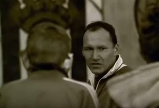 Zdjęcie przedstawia trenera głównego bohatera - dojrzałego mężczyznę, który stoi przed dwoma chłopcami i mówi do nich. Ma krótkie, ciemne włosy z zakolami oraz sportową bluzę.