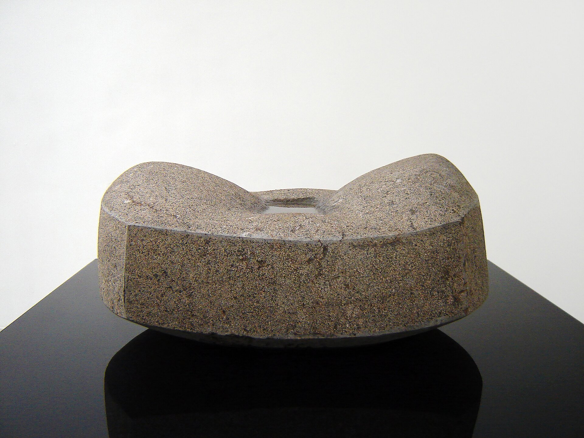 Ilustracja przedstawia rzeźbę „Źródło III”, autorstwa Macieja Wierzbickiego. Na zdjęciu ukazany jest kamienny, nieregularny kształt płaskiego sześcianu z małym prostokątnym wgłębieniem pośrodku, w którym znajduje się woda. Obiekt ustawiony jest na gładkim blacie czarnego granitu. Tło stanowi biała płaszczyzna.