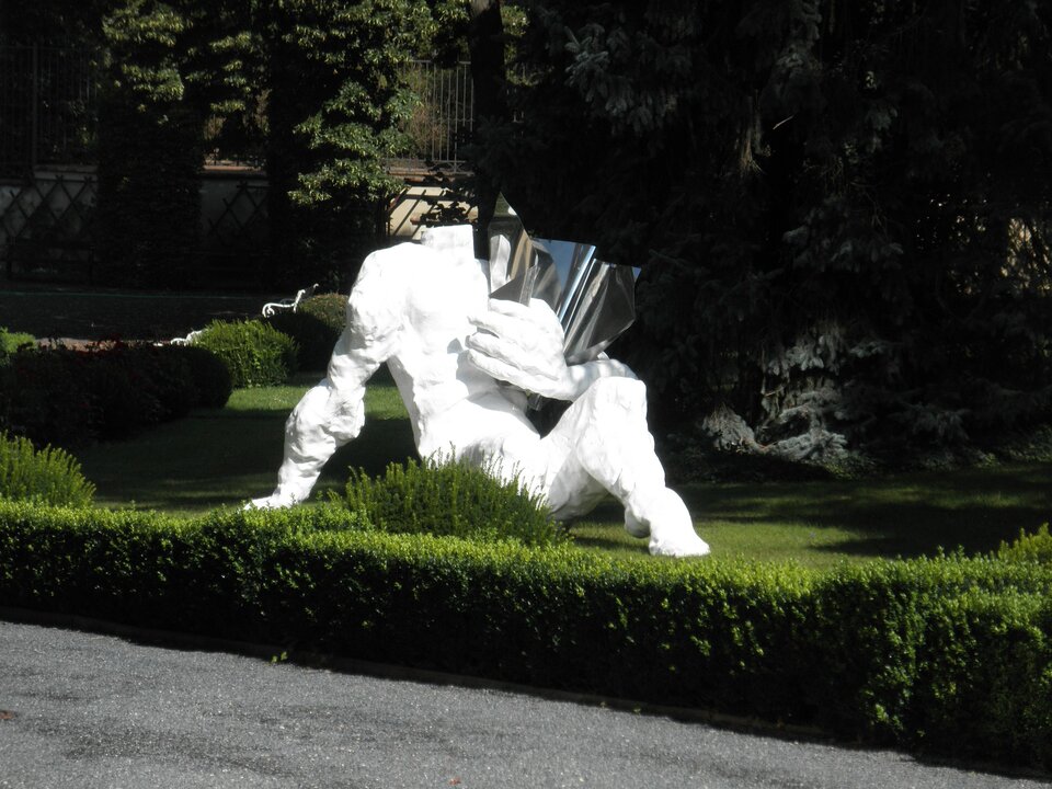 Zdjęcie przedstawia dzieło autorstwa Tomasza Koclęgi, pod tytułem: Rzeźba figuratywna. Na fotografii znajduje się biała rzeźba, przedstawiająca postać bez głowy. W jej lewej ręce znajduje się srebrny przedmiot, przypominający zwinięty arkusz. Postać siedzi na trawie z ręką opartą o ziemię. Rzeźba umieszczona jest w plenerze, gdzie rosną zielone krzewy oraz drzewa.