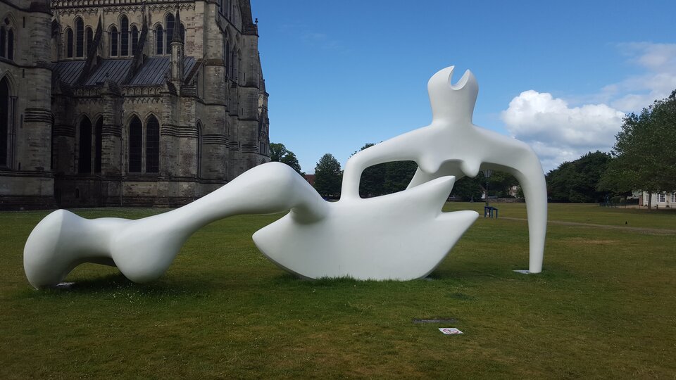 Fotografia przedstawia rzeźbę Henry'ego Moore'a ustawioną przed katedrą w Salisbury. Rzeźba ukazuje leżącą postać kobiecą w abstrakcyjnej formie, o płynnych kształtach.  Jej pozycja – z jedną nogą wyciągniętą, drugą ugiętą oraz jedną ręką wspierającą tułów – sugeruje zrelaksowanie, swobodę.  Głowa postaci jest uproszczona, a ciało zdeformowane, zredukowane, o płynnej linii i abstrakcyjnym kształcie.  Rzeźba znajduje się w otwartym krajobrazie, a jej jasna, gładka powierzchnia kontrastuje z trawiastym podłożem. Dzięki swojej lekkiej, asymetrycznej konstrukcji wydaje się jednocześnie stabilna i dynamiczna.