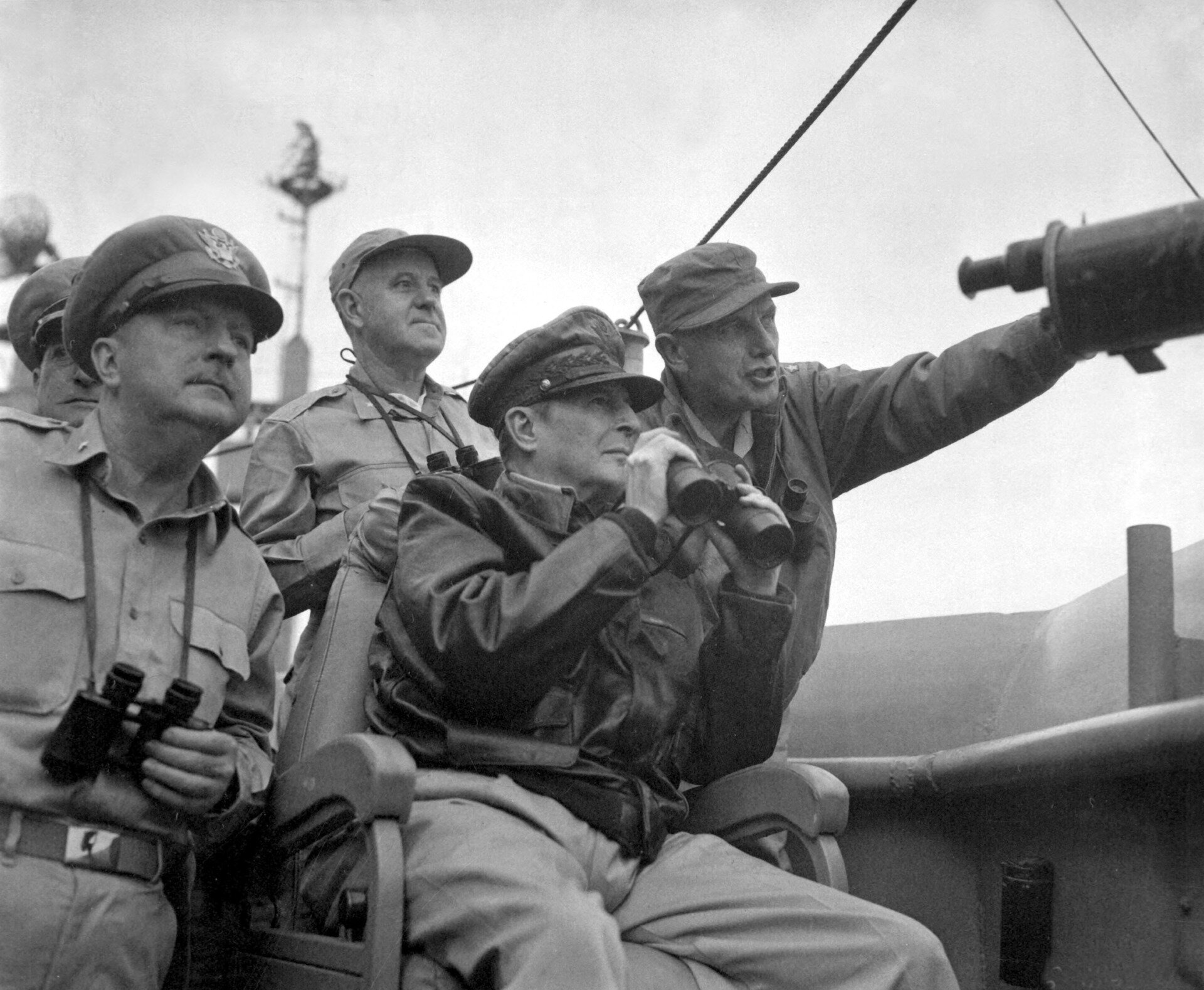 Fotografia czarno-biała przedstawiająca siły ONZ (amerykańskie) , którymi dowodził generał Douglas MacArthur. Dowódca siedzi na krześle,  w rękach trzyma lornetkę. Każdy z obecnych przy nim obserwujący również ma swoją lornetkę, jeden z nich wskazuje ręką kierunek w którym należy patrzeć. 