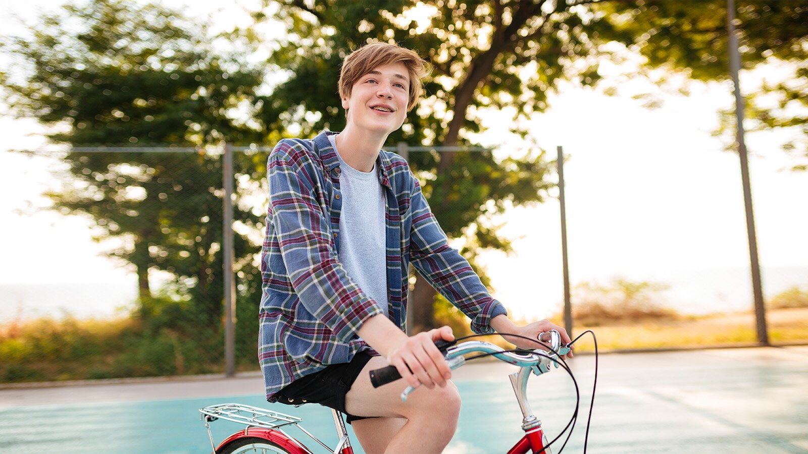 Zdjęcie przedstawia uśmiechniętego nastolatka, ubranego w krótkie, czarne spodenki, białą koszulkę i kraciastą koszulę, który siedzi na rowerze. Ręce trzyma na kierownicy roweru. W tle widoczne drzewa oświetlone słońcem.