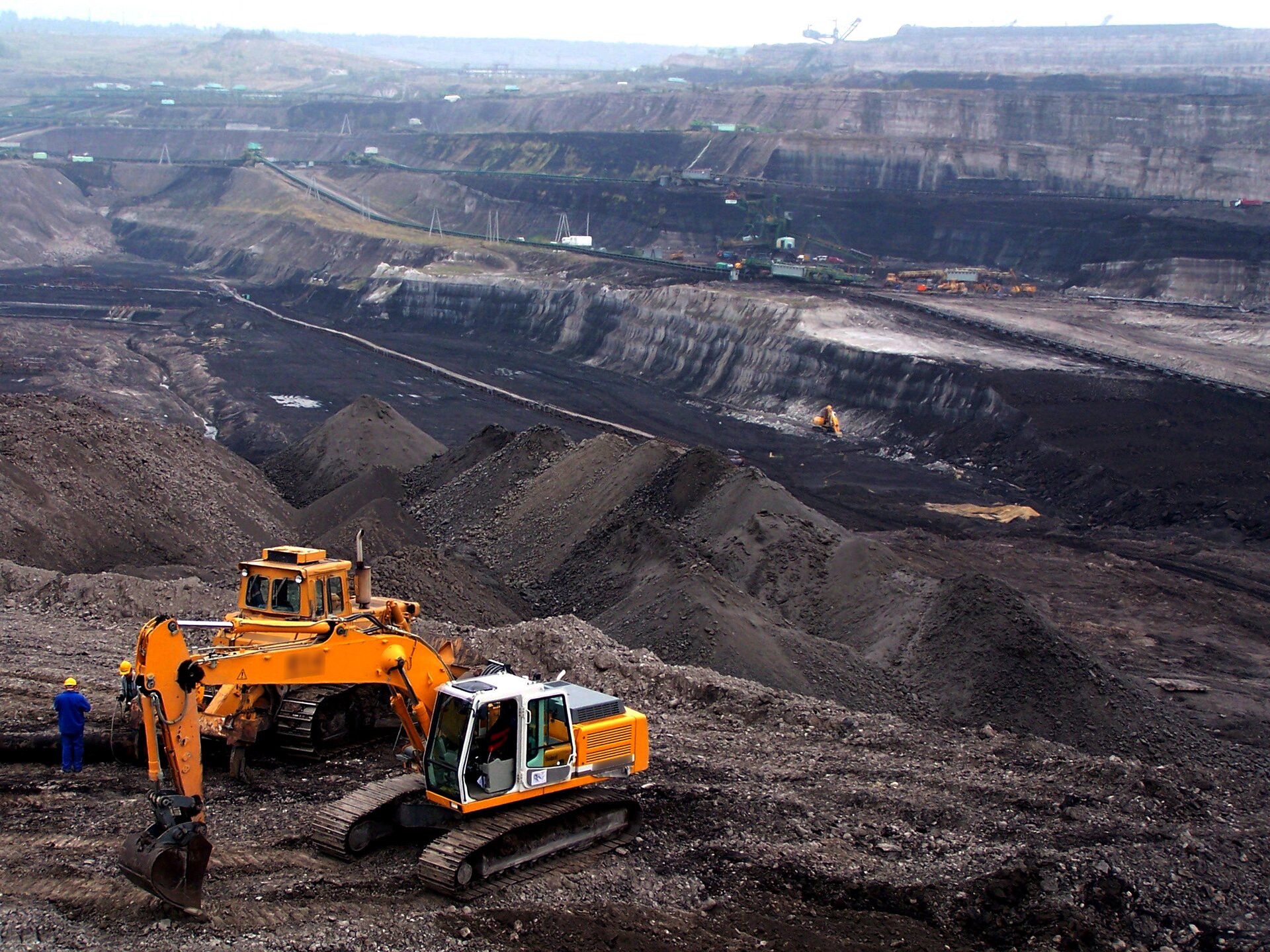 Na zdjęciu kopalnia odkrywkowa. Rozległe wyrobisko. Na pierwszym planie pomarańczowe koparki i pracownicy. Na drugim duże hałdy węgla. W tle kolejne maszyny. Kopalnie ma setki metrów średnicy, stanowi bardzo głębokie, jałowe wyrobisko w ziemi.