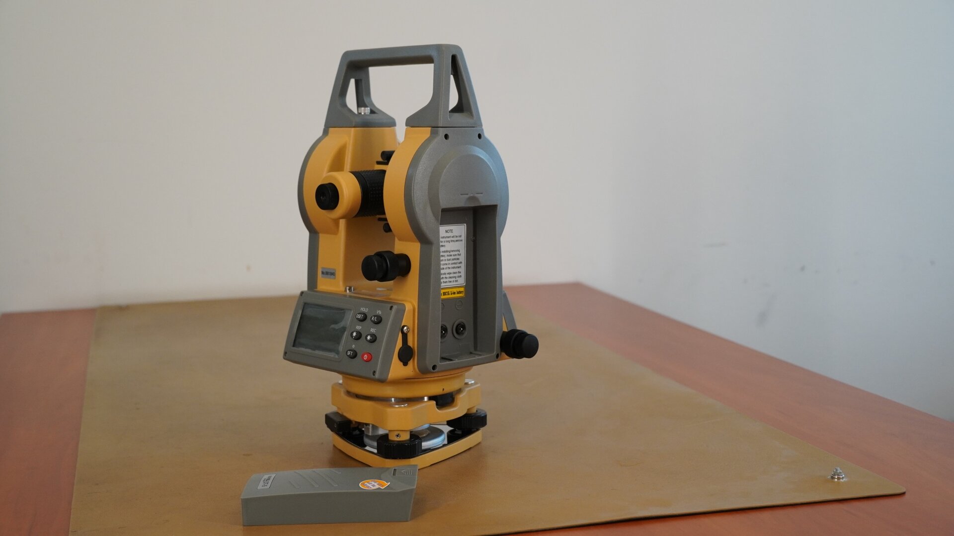 Zdjęcie przedstawia widok teodolitu z bocznej perspektywy. Widać na nim miejsce, w którym umieszcza się baterie. Zdjęto z niego osłonę, a baterię umieszczono na blacie przed instrumentem. Po lewej stronie widoczny jest okular, a pod nim wyświetlacz z przyciskami.