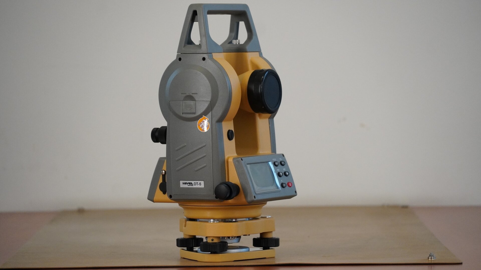 Zdjęcie ukazuje boczne ujęcie teodolitu. Po prawej stronie widoczny jest obiektyw, a poniżej wyświetlacz z przyciskami. Urządzenie posiada trójkątną podstawę w formie płytki sprężynującej. W każdym jej rogu znajduje się śruba poziomująca. Na śrubach osadzona jest spodarka, czyli podstawa instrumentu. W spodarce mieści się śruba dociskowa, a na spodarce okrągła alidada, czyli korpus urządzenia. Na nim zaś znajdują się pokrętła leniwki alidady i leniwki lunety oraz zacisk alidady i lunety. Konstrukcję instrumentu uzupełnia prostokątna rama z uchwytem w górnej części.