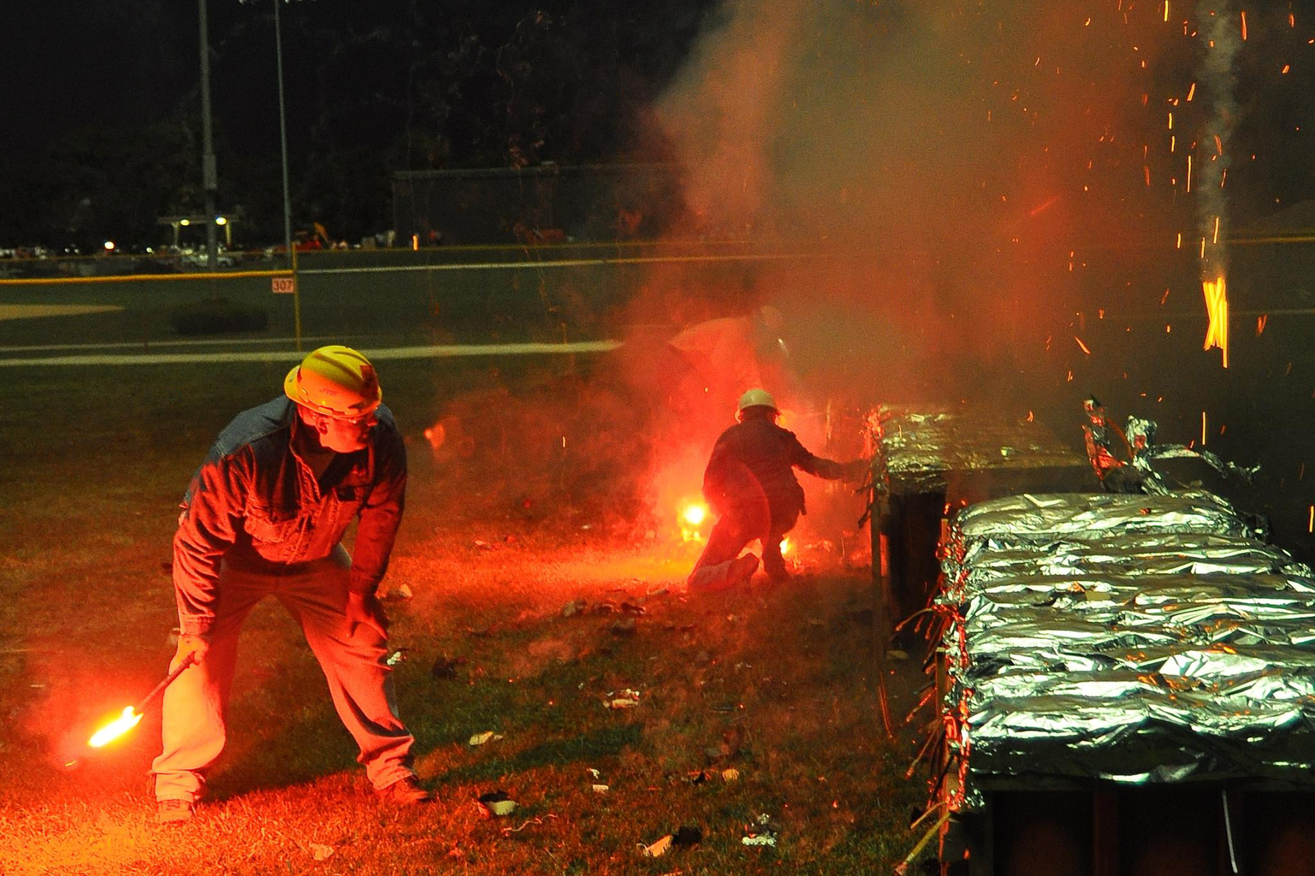 Zdjęcie przedstawia dwóch pracowników firmy pirotechnicznej przygotowujących profesjonalny pokaz sztucznych ogni. Obaj mają na sobie ubrania ochronne, kaski i trzymają w rękach zapalone flary, które posłużą im do podpalenia lontów.