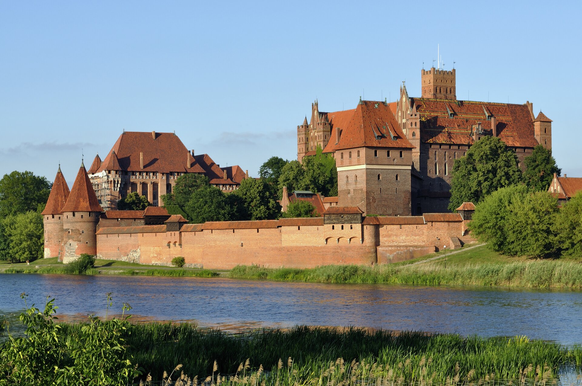 Na zdjęciu rozbudowany zamek z czerwonej cegły, otoczony murem, położony nad rzeką.