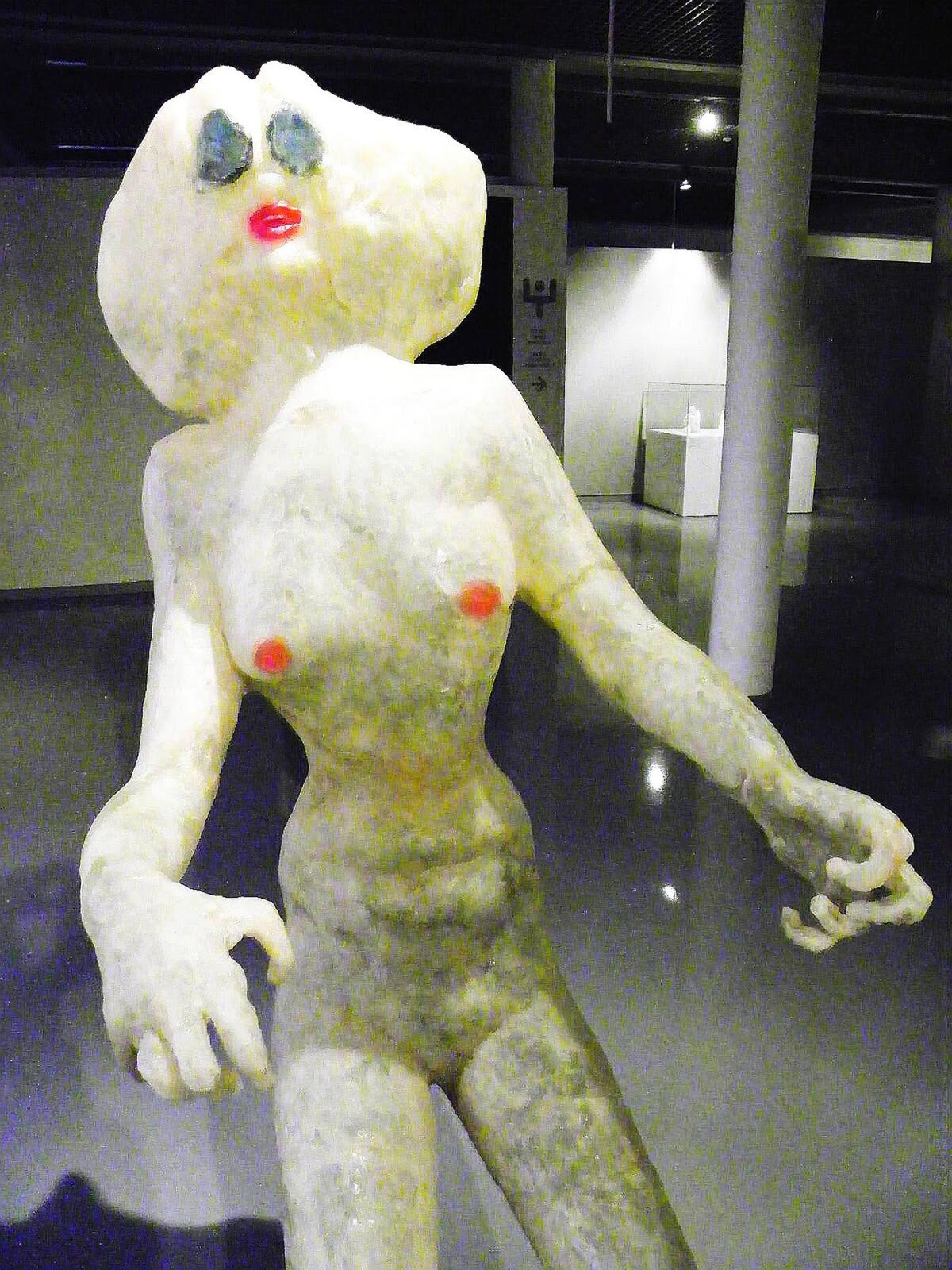 Zdjęcie przedstawia rzeźbę Aliny Szapocznikow Podróż.Naga postać kobieca w jasnym kolorze, Ręce ma wyciągnięte przed siebie, palce zgięte w nienaturalny sposób. Sutki piersi zaznaczone są na czerwone. Głowa przypomina niekształtna kulę, jak głowa bałwana, z czerwonymi ustami i niebieskimi dużymi oczami w jajowatym kształcie.