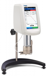 Zdjęcie przedstawia reometr typu stożek. Urządzenie zbudowane jest z panelu z wyświetlaczem osadzonym na regulowanym wieszaku, wspartym na półokrągłym stelażu. Pod skrzynką urządzenia znajduje się cylindryczny, metalowy element.