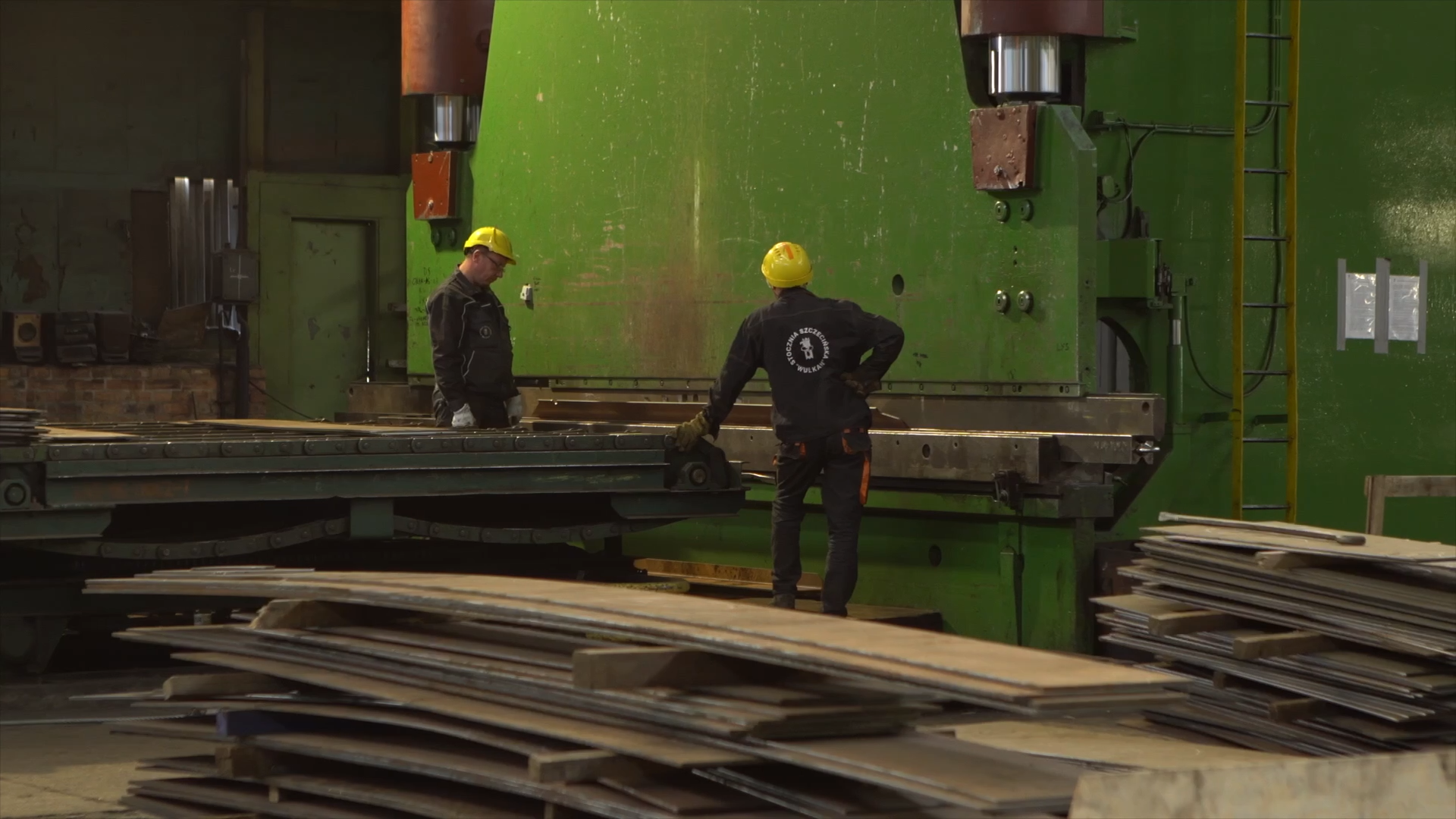 Zdjęcie przedstawia prasę krawędziową w stoczni. Stoi przy niej dwóch pracowników w strojach roboczych i kaskach. Prasa to duże prostokątne urządzenie służące do gięcia na przykład blach.