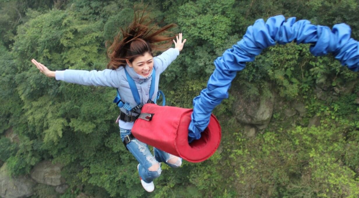 Skok na bungee Zdjęcie przedstawia na tle zielonej roślinności dziewczynę z rozpostartymi ramionami, która jest połączona z grubą niebieską pomarszczoną rurą z czerwonym pojemnikiem w kształcie walca. Włosy dziewczyny uniesione do góry.