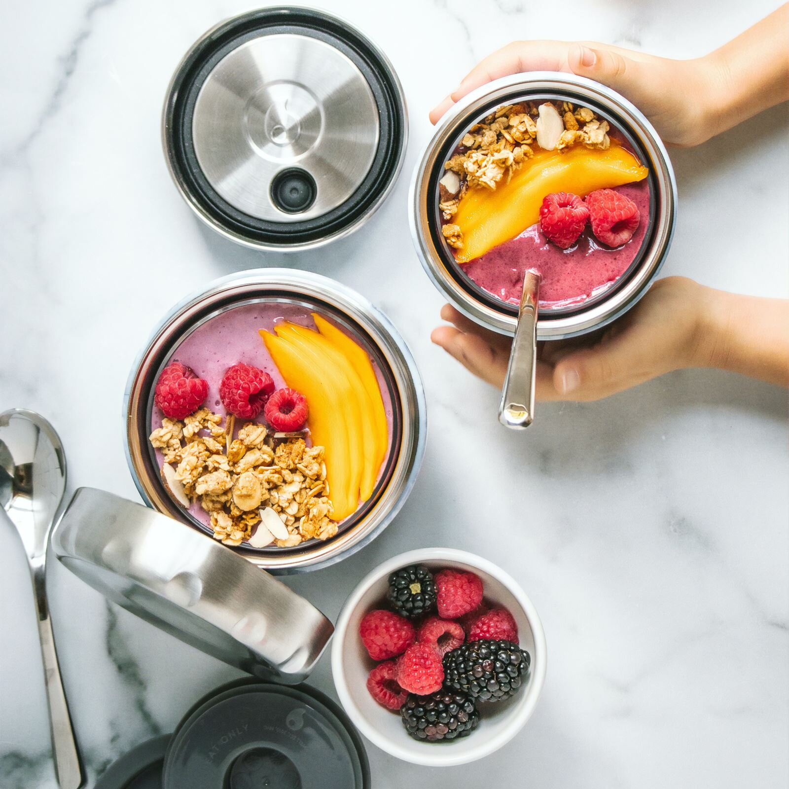 Zdjęcie przedstawia ustawione na blacie okrągłe metalowe boksy podobne do słoika a w nich widoczny jest różowy jogurt oraz owoce i płatki owsiane. Mamy pokrojone w plasterki mango i maliny. Obok stoi miseczka z owocami gdzie widoczne są jeżyny i maliny. Jeden boks trzyma ktoś w dłoniach.