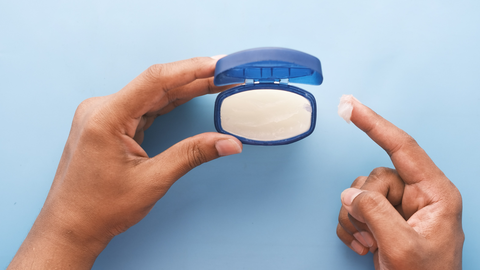 Zdjęcie przedstawia pojemnik z maścią trzymany przez kogoś w ręku. Na wskazującym palcu prawej dłoni widoczna jest nabrana z pojemnika maść.