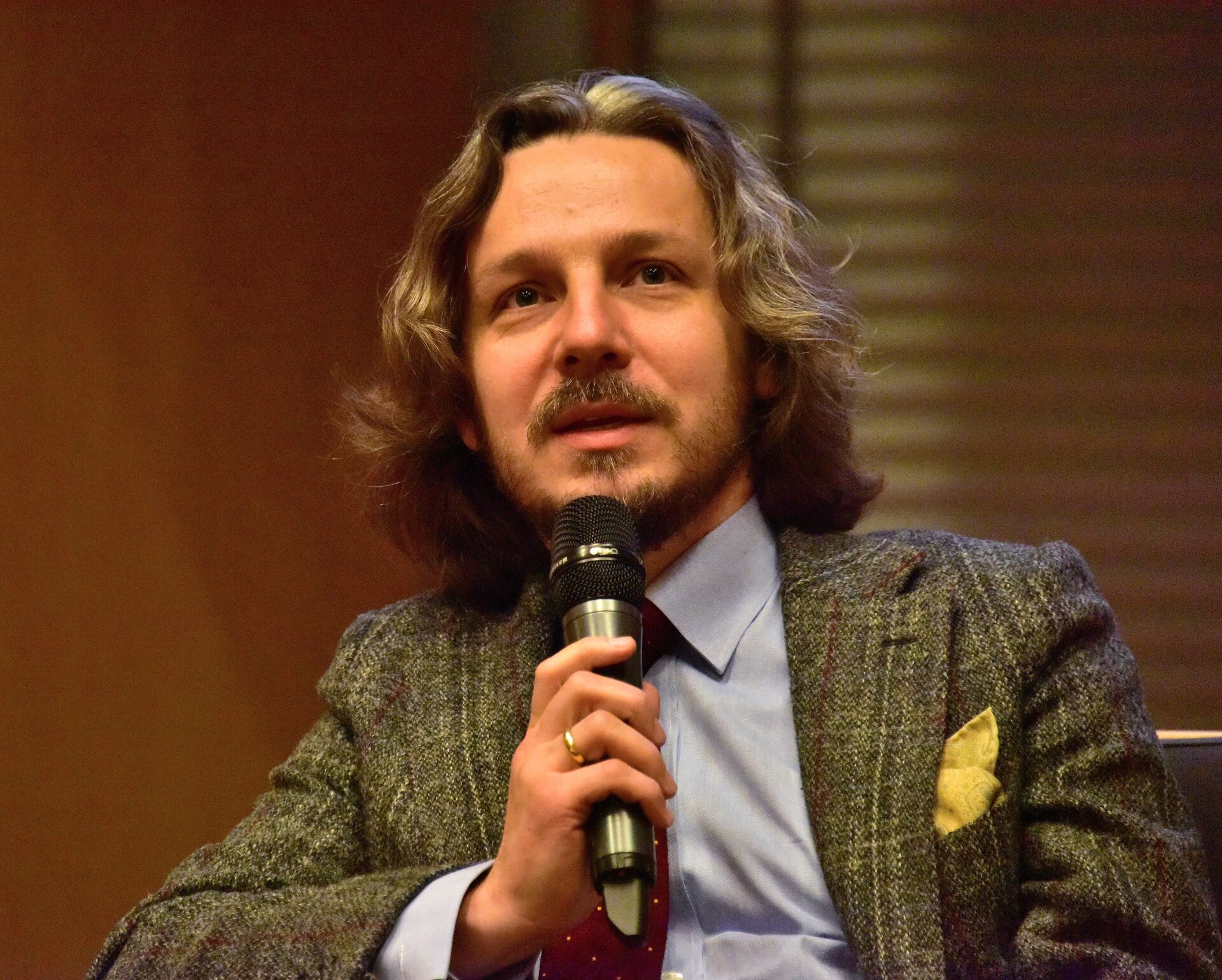 Fotografia przedstawiająca mężczyznę w średnim wieku. Jest to szatyn o lekko falowanych włosach do ramion. Ma szczupłą twarz, ciemne oczy, delikatnie zarysowaną brodę oraz wąsy. Ubrany jest on w marynarkę, spod której widać jasnoniebieską koszulę. W prawej górnej kieszeni marynarki wetknięta została żółta poszetka.