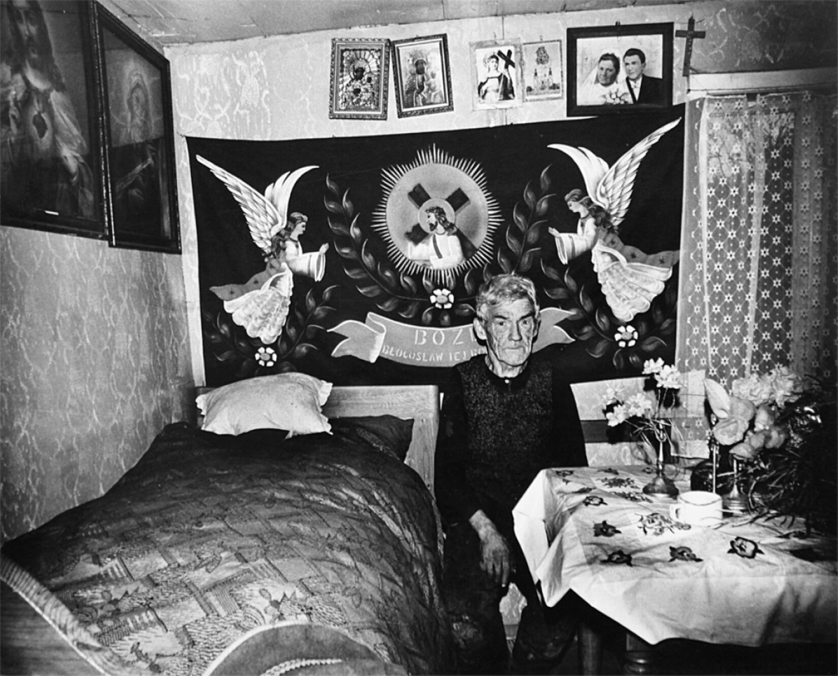 Czarno białe zdjęcie przedstawia mężczyznę w podeszłym wieku, o krótkich siwych włosach zaczesanych na bok. Ubrany jest w sweter, wełnianą kamizelkę oraz długie spodnie. Na jego twarzy widoczne są zmarszczki. Mężczyzna siedzi w pokoju na krześle, pomiędzy łóżkiem a stolikiem. Na ścianach pomieszczenia wisi  krzyż, obraz, makatka oraz zdjęcie ślubne. Na stoliku z jego lewej strony widać kwiaty w wazonach, a niewielkie okno znajdujące się po prawej stronie zasłonięte jest białą firanką. Tapeta widoczna na ścianie po prawej stronie jest jasna, w nieregularne wzory.  