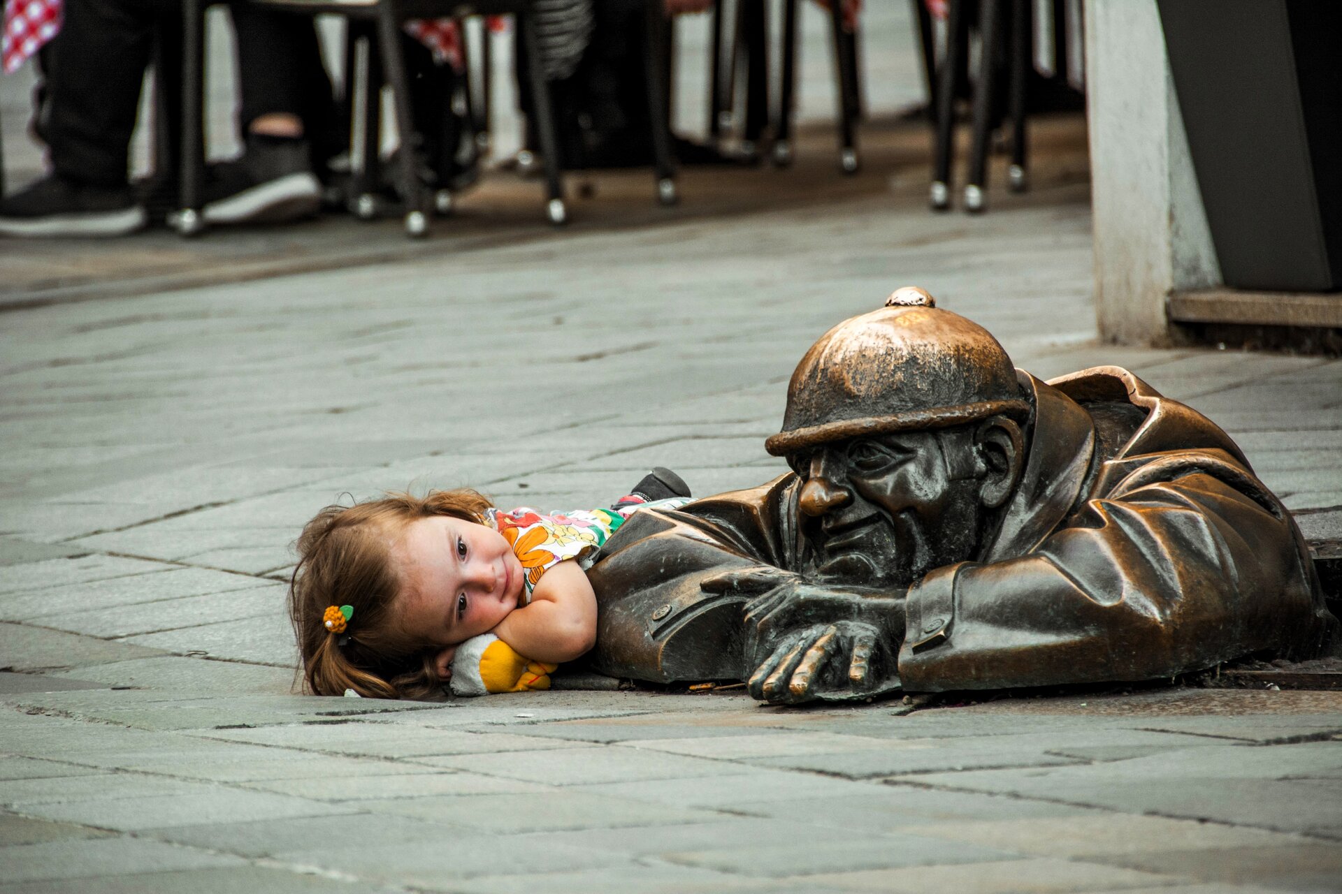 Zdjęcie przedstawia sytuację, w której mała dziewczynka leży na chodniku, przypatrując się rzeźbie z brązu. Rzeźba przedstawia robotnika, który wychyla się z kanału, opiera ręce na chodniku i z uśmiechem marzy. Scenkę obserwują ludzie z oddali.