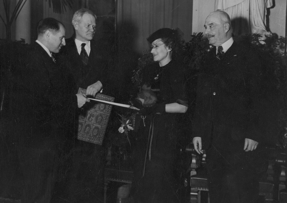 Zdjęcie przedstawia cztery osoby. Kobietę i trzech mężczyzn. Od lewej strony stoi dwóch mężczyzn w średnim wieku. Są ubrani w garnitury. Jeden z nich wręcza kobiecie nagrodę. Kobieta jest w średnim wieku. Ubrana w suknię, toczek z niewielkim welonem i rękawiczki. Skierowana jest twarzą do mężczyzny trzymającego nagrodę. Obok kobiety, po prawej stronie zdjęcia, stoi starszy mężczyzna. Jest ubrany w garnitur.