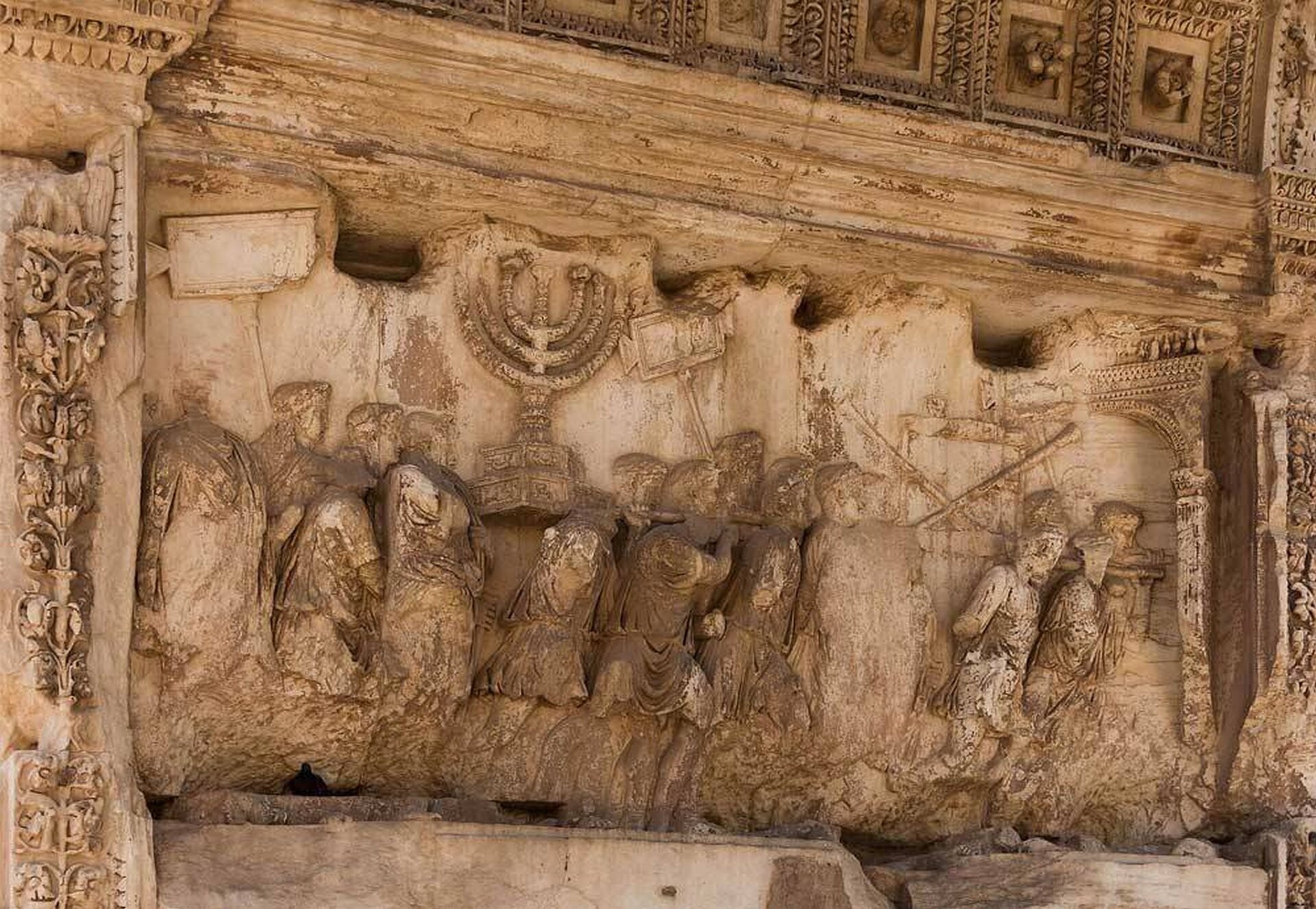 Ilustracja przedstawia fragment łuku tryumfalnego Tytusa, uświetniającego zdobycie przez cesarza Jerozolimy. Na fotografii widoczny jest detal ukazujący pochód triumfalny. Żołnierze rzymscy niosą zdobycze wojenne, m.in. duży siedmioramienny świecznik.