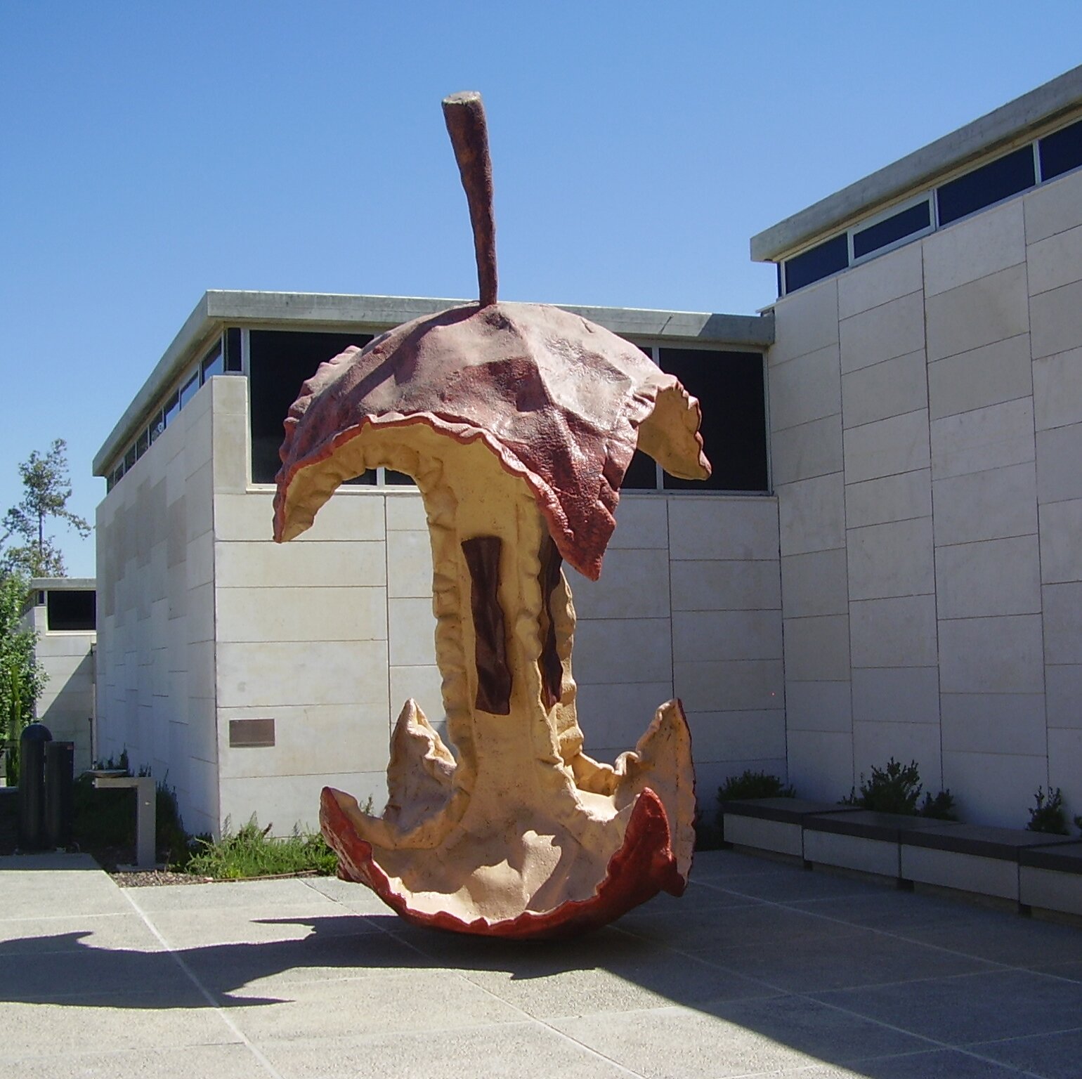 Zdjęcie przedstawia dużą rzeźbę ogryzka jabłka, w oddali widać budynek w kształcie prostokąta.