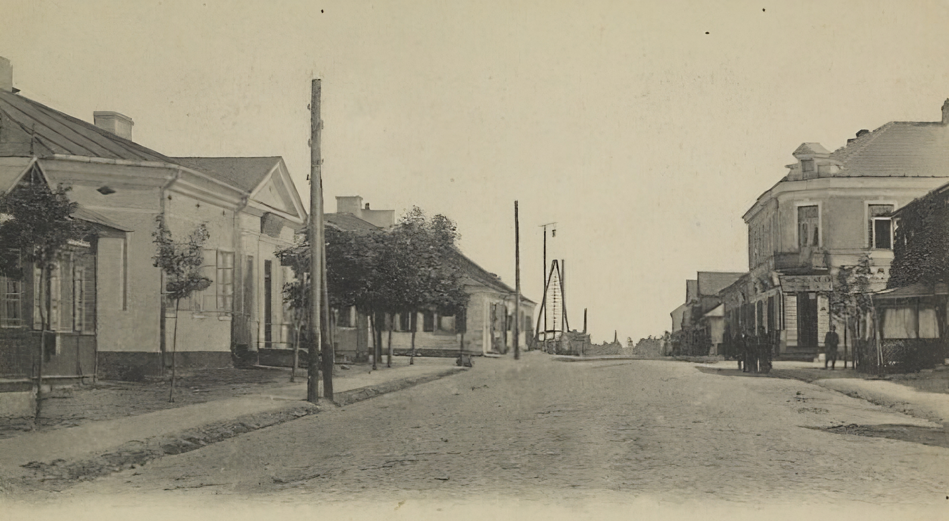 Kliknij, aby powiększyć Zdjęcie przedstawia brukowaną ulicę z chodnikami dla pieszych. Przy chodniku po lewej stronie ustawione są słupy elektryczne po stronie ulicy. Od strony budynków obsadzony jest drzewami. Najbliższy budynek jest drewniany. Dalej znajduje się murowany budynek, który przylega od północy do prostopadłej ulicy. Na rogu tej ulicy po prawej stronie znajduje się piętrowy gmach. Na pierwszym piętrze tego budynku jest narożny balkon. Na parterze znajdują się sklepy. Przed budynkiem stoją ludzie. W dali widać po obu stronach ulicy drewniane domy nakryte dwuspadzistym dachem.