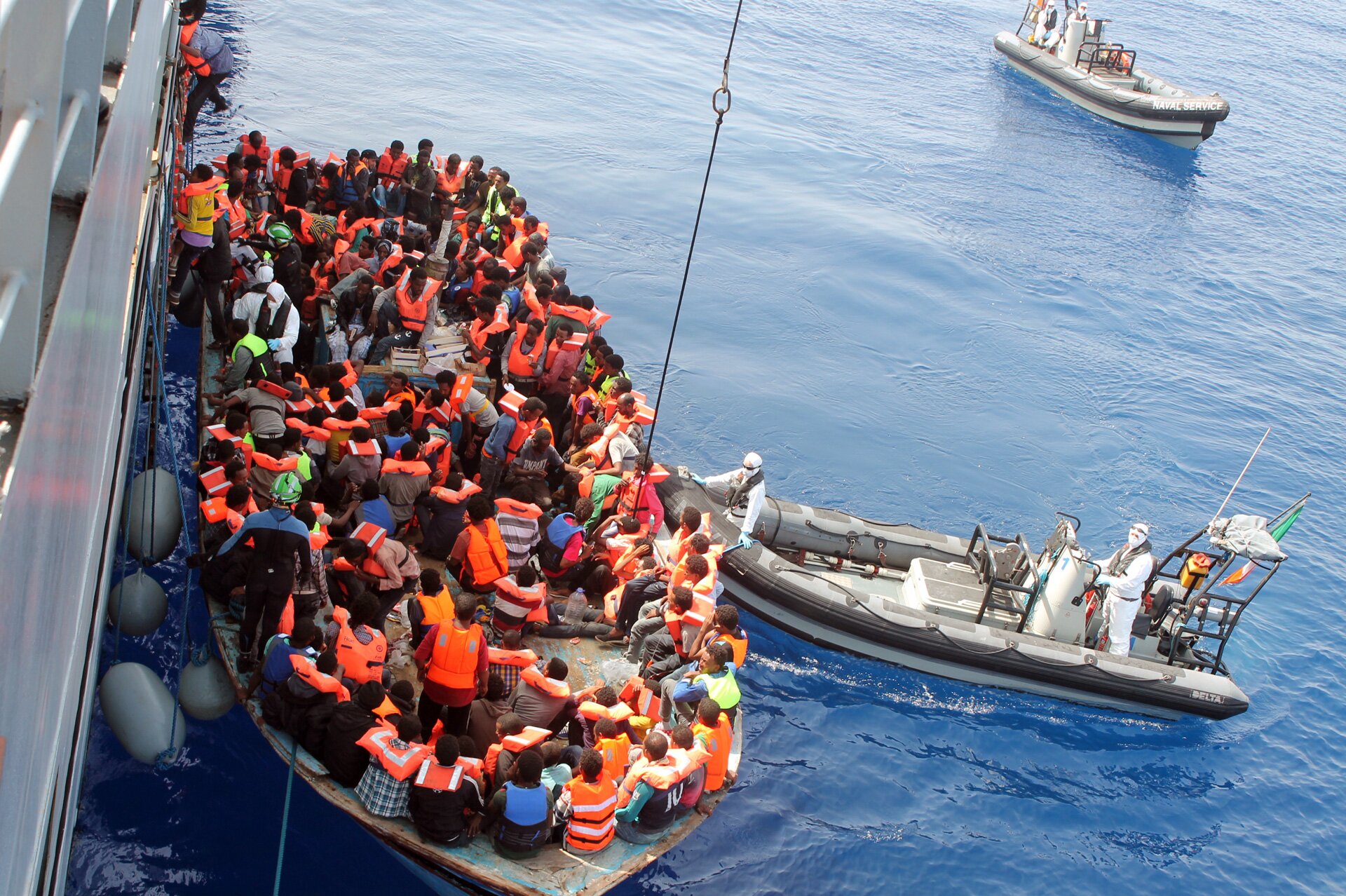 Na zdjęciu są uchodźcy w kapokach na łodzi ratunkowej u wybrzeży Włoch. Łódź ratunkowa znajduje się bezpośrednio przy bocznej ścianie ogromnego statku. Kilku z uchodźców jest w trakcie wsiadania na statek, wspinając się po drabince. Obok łodzi ratunkowej znajduje się motorówka z dwoma pracownikami w kombinezonach ochronnych na niej. W tle widoczne jest morze i druga motorówka.