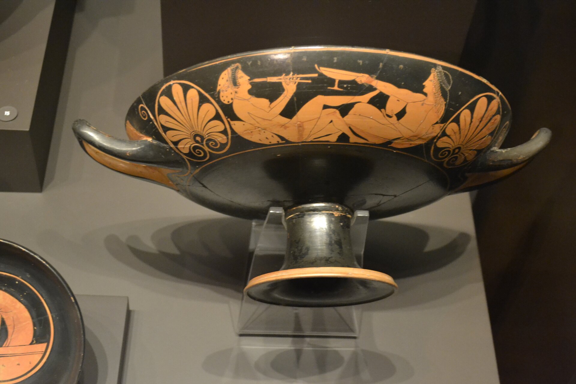 Fotografia autorstwa Jerónimo Roure Pérez przedstawia starożytny kyliks pochodzący z Attyki. Naczynie widoczne jest od dołu, ponieważ jest zawieszone na ścianie ekspozycyjnej. Naczynie ma wąską stopę, która rozszerza się. Po obu stronach wazy znajdują się uszy, które mogły służyć do przenoszenia naczynia lub pełnić funkcję ozdobną. Na powierzchni naczynia namalowana została para muzyków grająca na aulosach. Obok muzyków umieszczono ornamenty, kształtem przypominające liście. Waza została wykonana w stylu czerwonofigurowym.