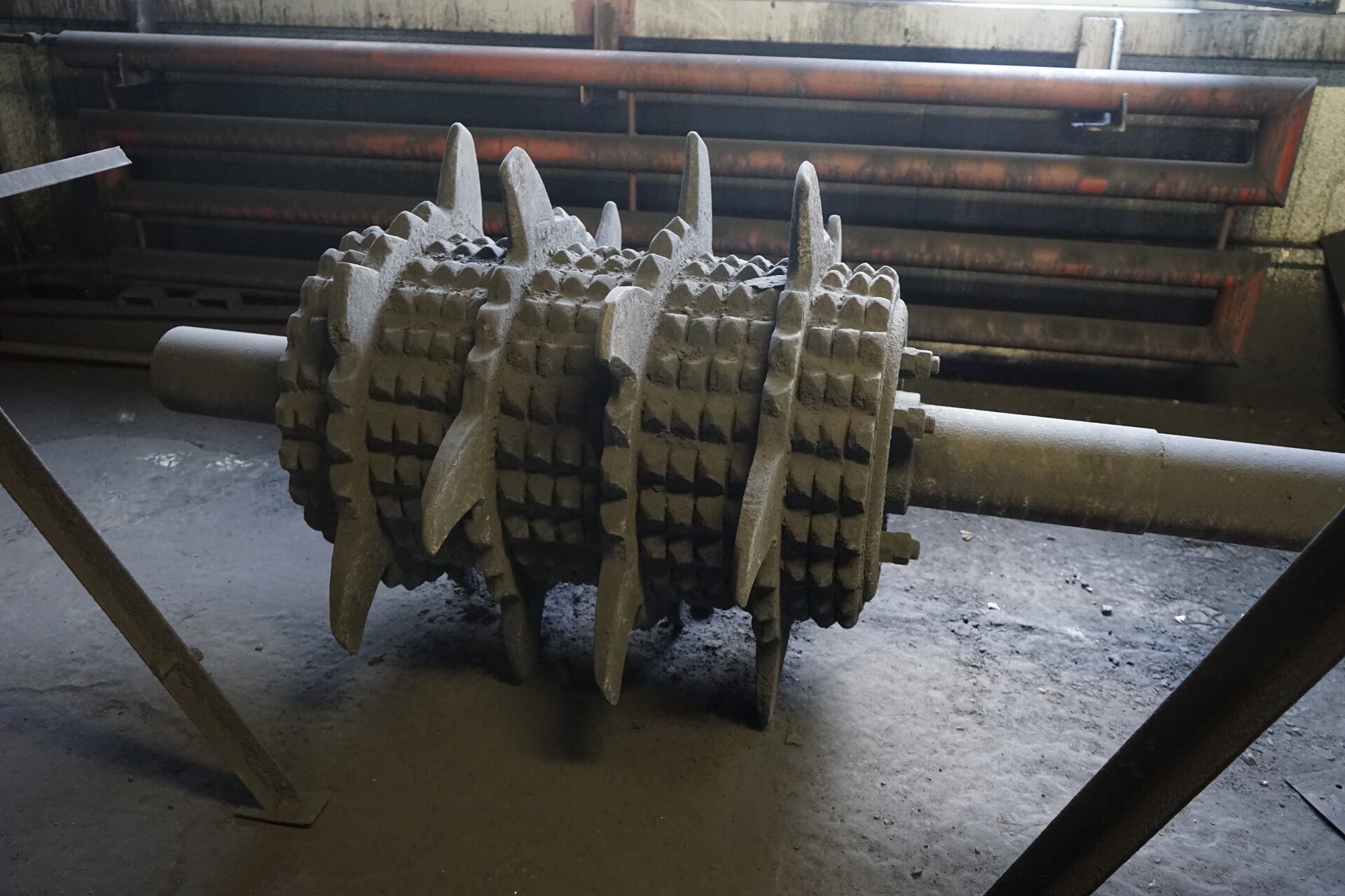 Zdjęcie przedstawia walec stanowiący element budowy małej kruszarki walcowej. Walec osadzony jest na długim wale. Powierzchnia walca jest uzębiona. To znaczy posiada wypustki oraz rozmieszczone w czterech pasmach zaostrzone, lekko wygięte w łuk blaszki. Poniżej zdjęcia widoczny jest przycisk multimedialny z nagraniem audio. Treść nagrania jest zgodna z tytułem zdjęcia.