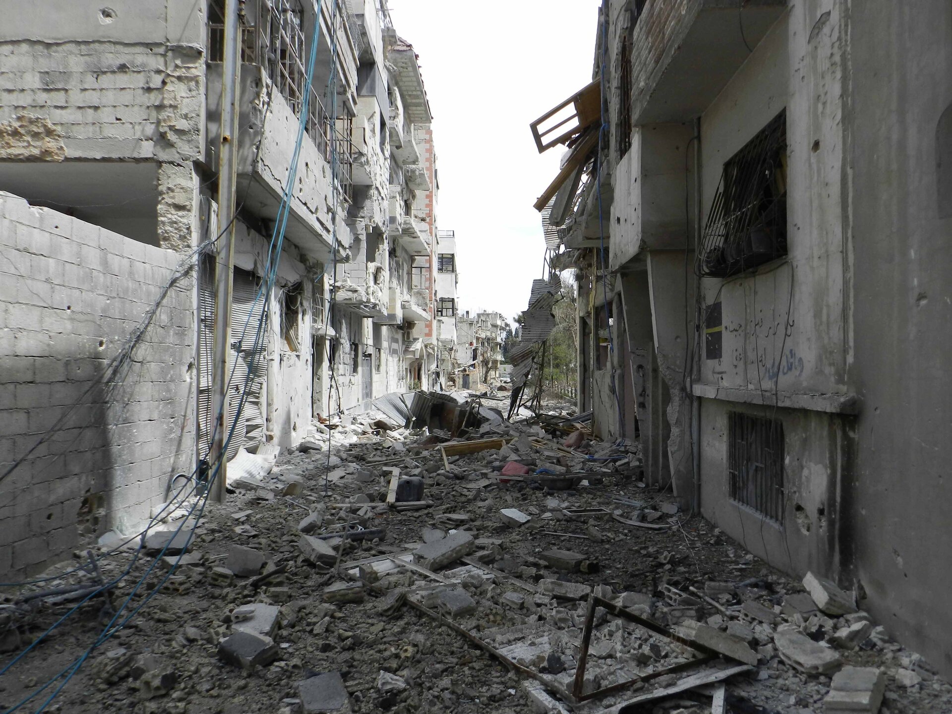 Zdjęcie, które przedstawia zniszczenia w Hims wywołane ofensywą wojska syryjskiego. Widoczne ruiny, zniszczenia po działaniach wojennych.
Po prawej i lewej stronie widać zbombardowane domy. Nie ma w nich okien, drzwi. Widoczne żelazne kraty i zwisające kable z domów. Po prawej stronie część domu zupełnie zniszczona. Widoczne pustaki na domach. Pomiędzy domami ulica, na której leży sporo gruzu.