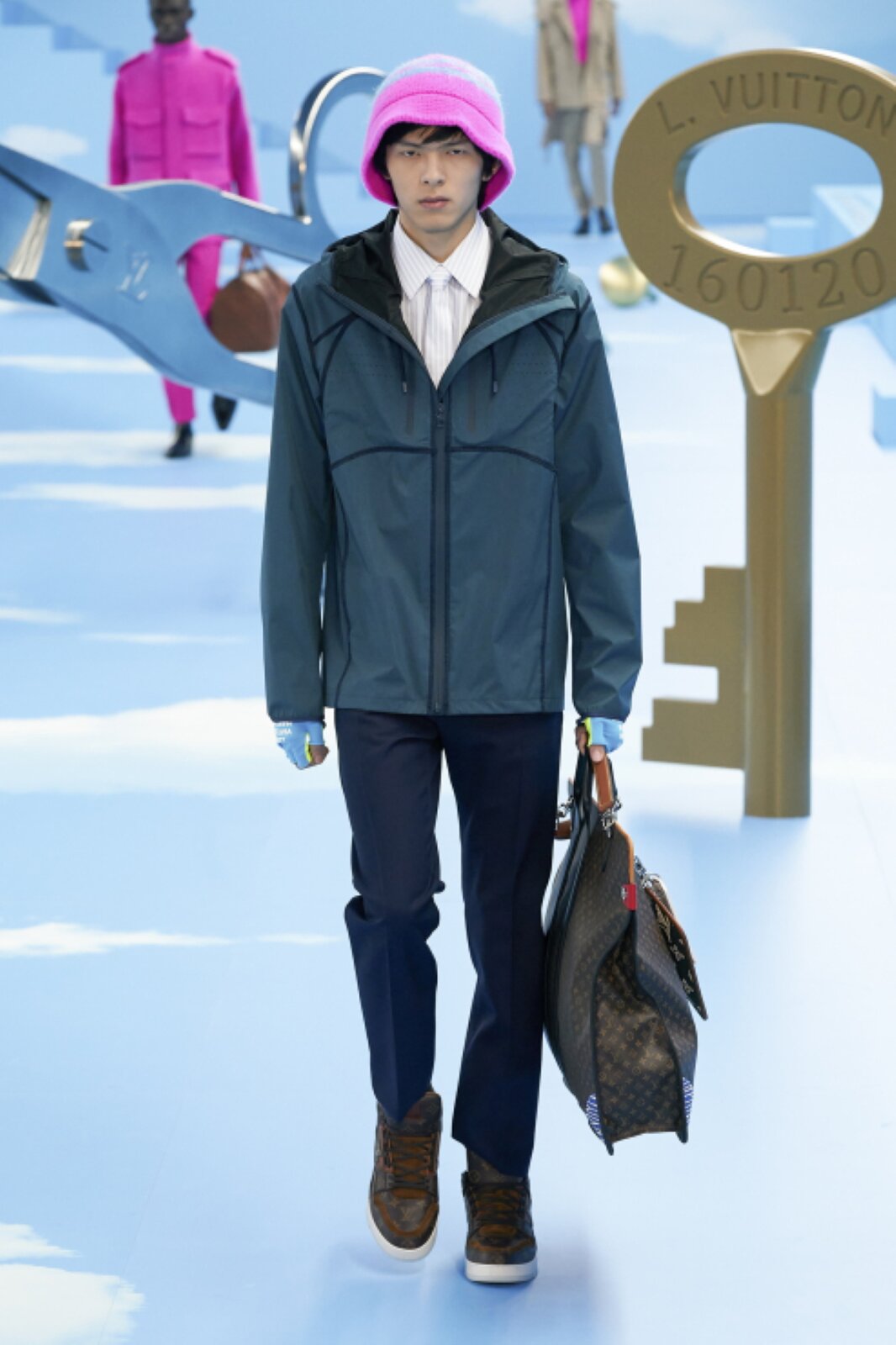 Fotografia przedstawia młodego mężczyznę, który ma na sobie niebieską rozpinaną kurtkę z czarnymi detalami, niebieskie rękawiczki odsłaniające palce, ciemnogranatowe spodnie wyprasowane na kant, białą koszulę, biały krawat oraz brązowe buty z białymi podeszwami. Na głowie ciemnowłosego mężczyzny widać jaskraworóżową czapkę w stylu „bucket hat”, a w jego lewej dłoni dużą, ciemną torbę w jasnobrązowe drobne wzory. 