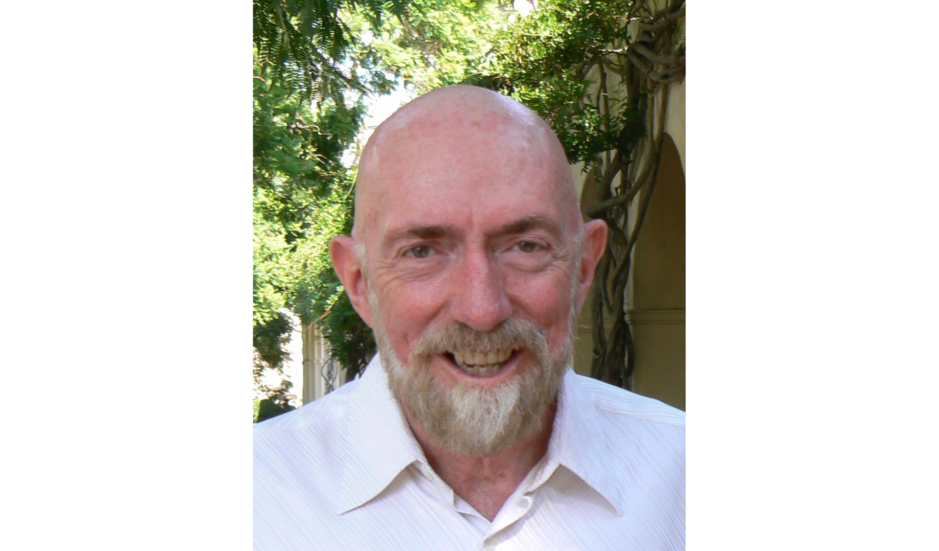 Zdjęcie przedstawia szeroko uśmiechniętego pana. Łysy, z odstającymi uszami. Siwa broda i wąsy.