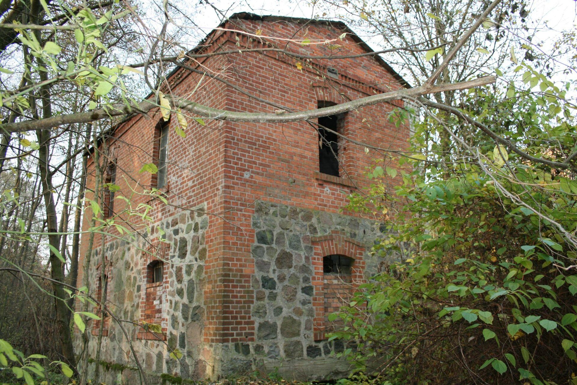 Zdjęcie przedstawia piętrowy, ceglany budynek na planie prostokąta. Okna na parterze są częściowo zamurowane, a ściany są wyłożone kamieniami na zmianę z czerwoną cegłą. Wokół budynku rośnie wiele drzew i krzewów. Niebo jest niebieskie, bezchmurne. Zdjęcie zostało wykonane w świetle dziennym.