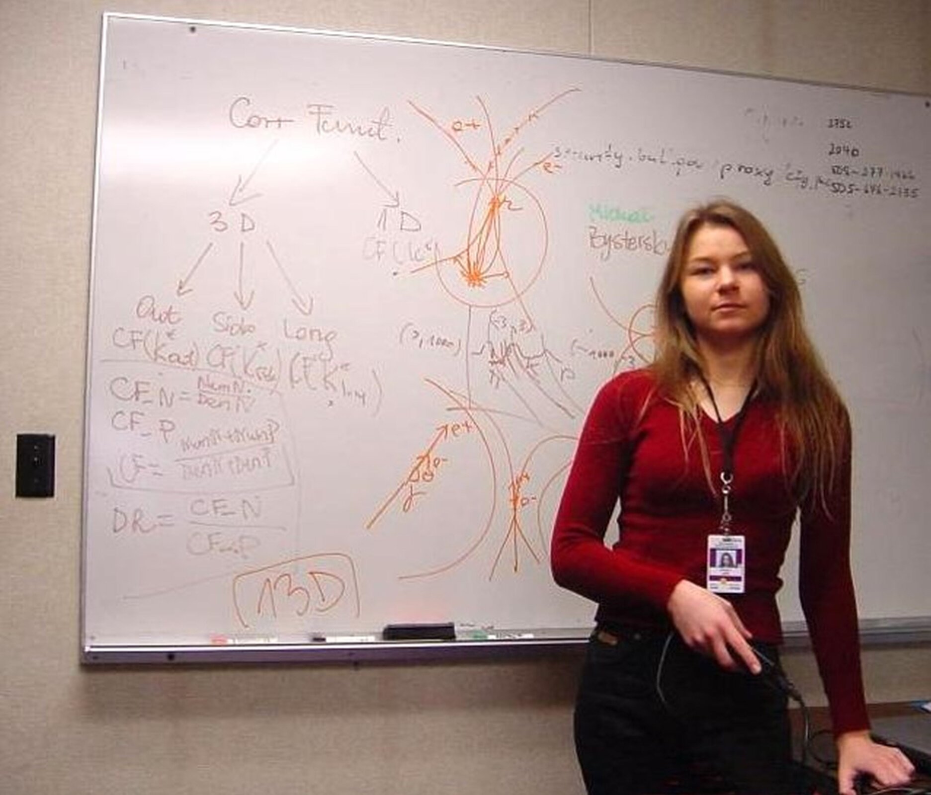 Rys. 4. Na zdjęciu widać młodą kobietę, za nią na ścianie widoczna biała tablica, na tablicy zapisano nieczytelnie wyrażenia najprawdopodobniej fizyczne, w centralnej części tablicy schematyczne rysunki.