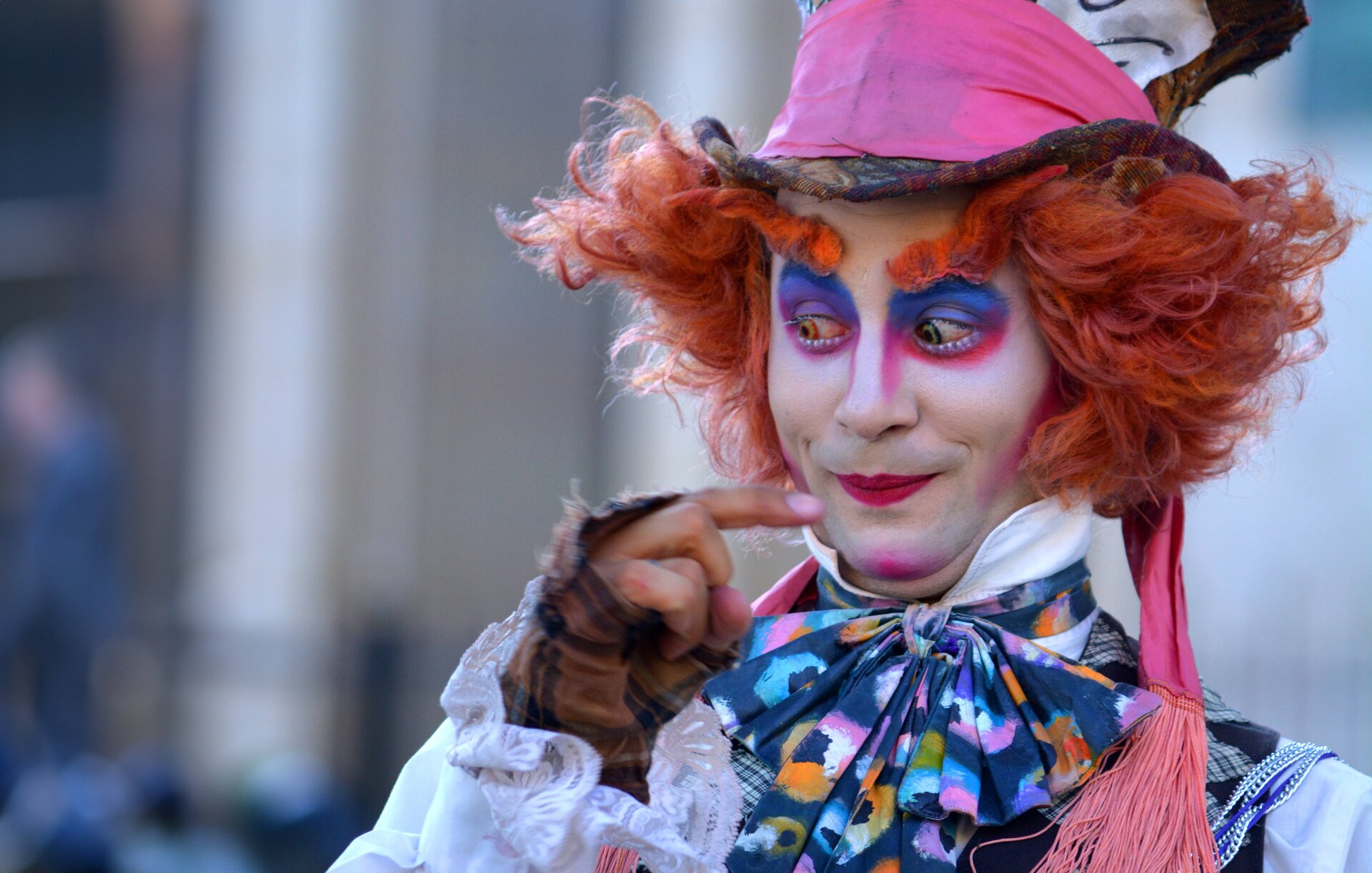 Zdjęcie przedstawia mężczyznę w pełnym makijażu ucharakteryzowanego na postać z bajki. Przypomina on klauna o białej twarzy i wieloma barwnymi odcieniami wokół oczu. Na głowie nosi perukę ze sterczącymi włosami. Nosi również strój w krzykliwych kolorach. Na głowie ma duży kapelusz przepasany brudną, różową szarfą.