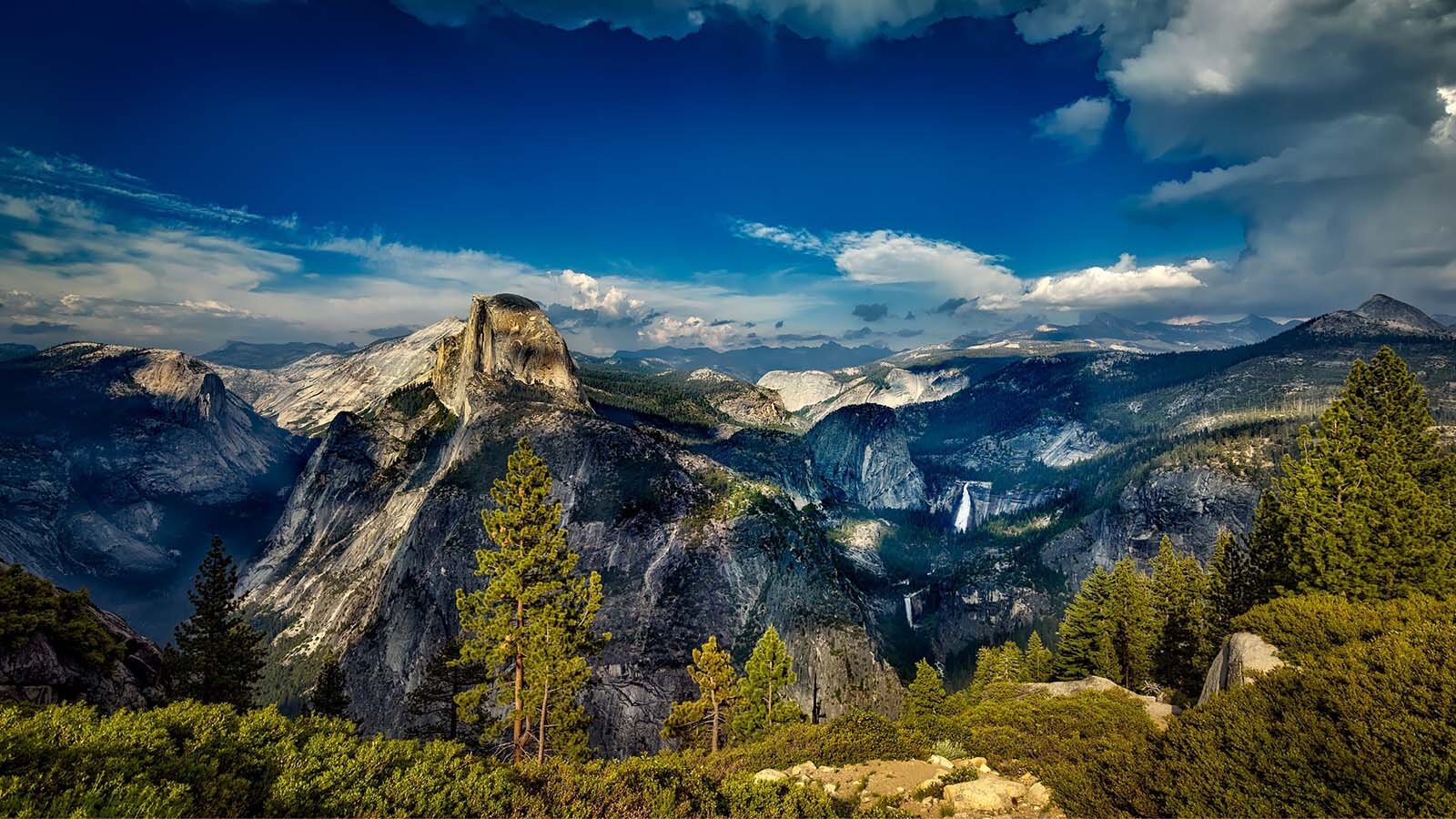 Zdjęcie przedstawia skaliste pasma górskie oświetlone promieniami słońca. Skały mają kolor od jasnoszarego po ciemnoszary i w dolnych partiach porośnięte są kępami iglastych drzew. Na pierwszym planie, u dołu, widoczne są pojedyncze ilaste drzewa oraz korony drzew. U góry widoczne jest intensywnie niebieskie niebo z pasami białych chmur nad szczytami.