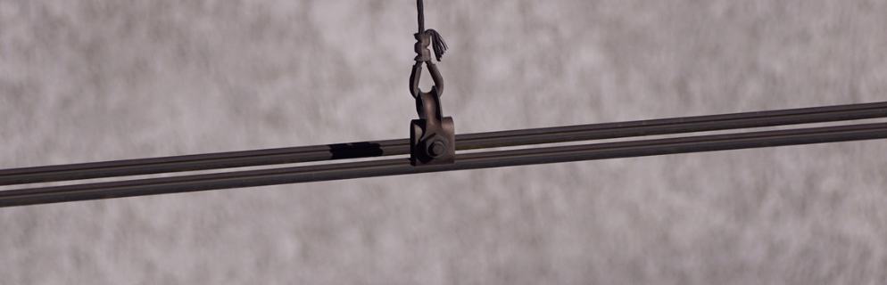 Zdjęcie przedstawia drut jezdny. Jest to prosty drut metalowy. Na zdjęciu widoczna jest także dolna końcówka wieszaka, służącego do zawieszenia drutu na linie nośnej. 