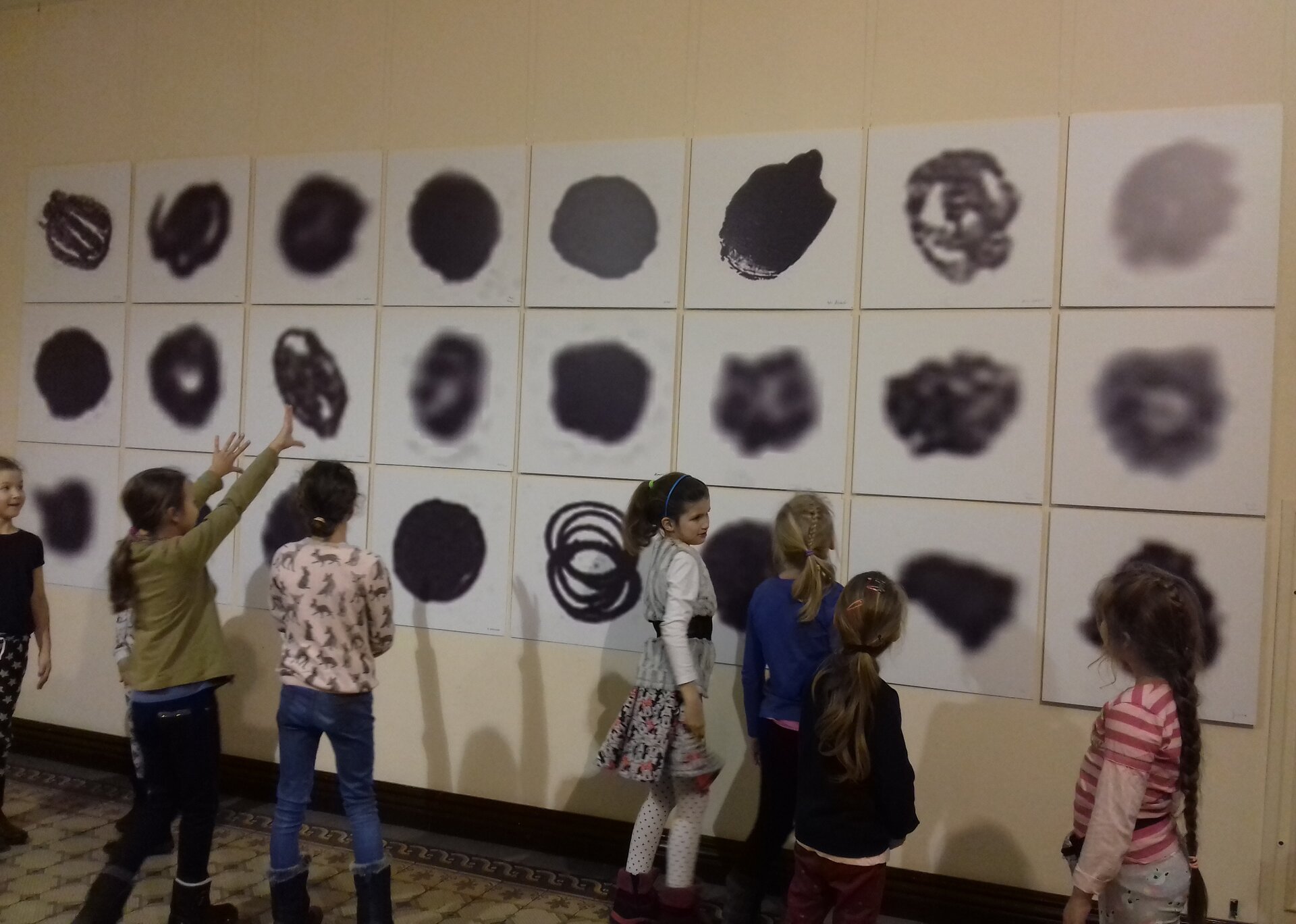 Ilustracja przedstawiająca dzieci oglądające wystawę sztuki pt.: "Punkty". Grupa dzieci stoi przed ścianą, na której znajdują się w trzech rzędach prace z narysowanymi czarnymi punktami. Jedne są kształtne, inne mają poszarpane brzegi. Każdy punkt ma inny kształt.