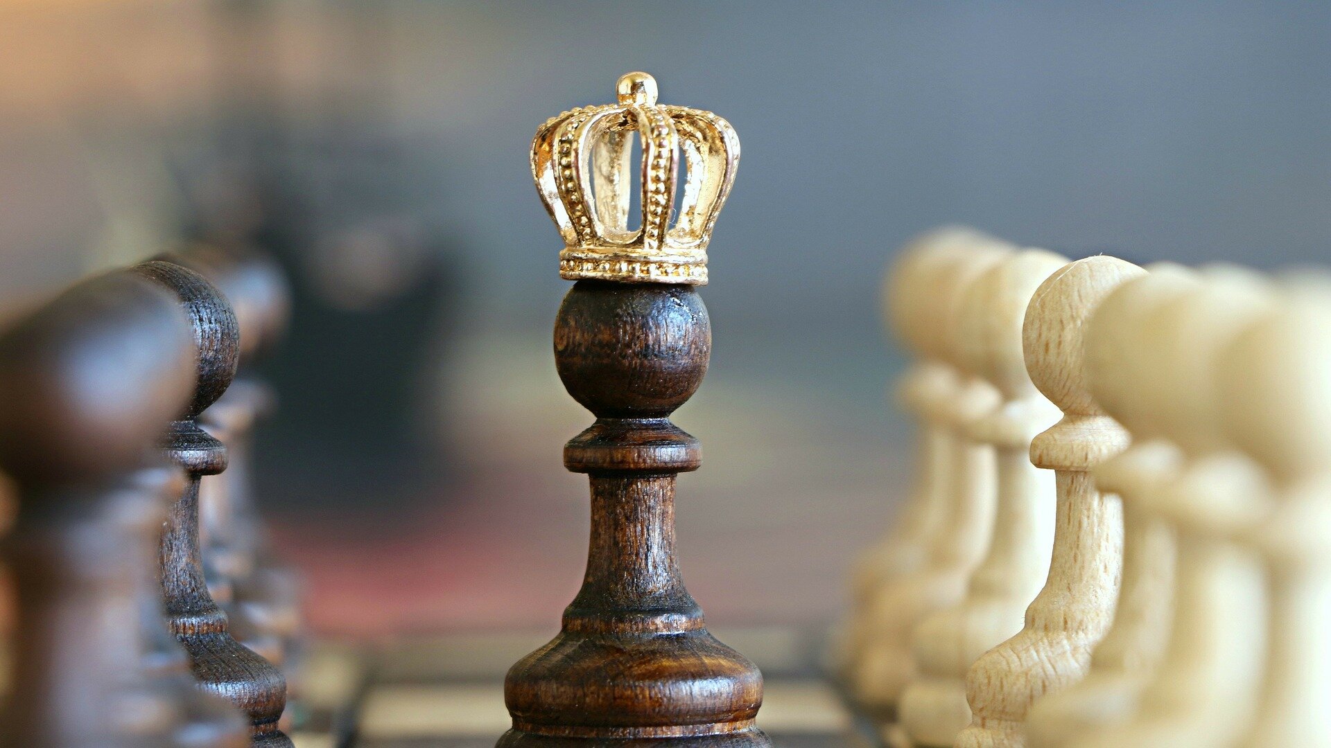 Zdjęcie przedstawia szachownicę, na której stoją naprzeciw siebie figury szachowe w białym i brązowym kolorze. Pomiędzy nimi znajduje się ciemna figura króla.
