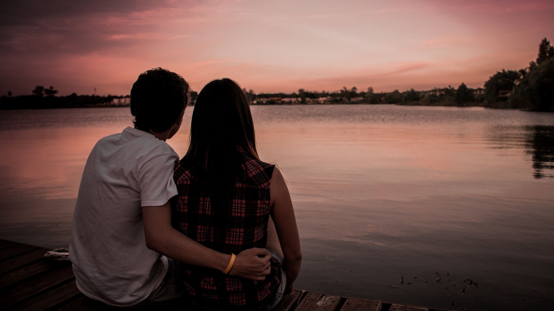 Zdjęcie przedstawię parę ludzi, którzy objęci siedzą nad wodą. Jest tuż po zachodzie słońca. Niebo jest jeszcze różowe, chociaż nie widać już słońca nad horyzontem. Jest tuż przed zapadnięciem zmroku. Jest ciepło, para ma lekkie ubrania z krótkimi rękawkami.