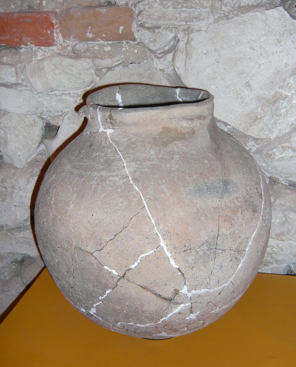 Na ilustracji ukazana jest duża gliniana amfora o kulistym kształcie, ustawiona na jasnobrązowym podłożu, na tle kamienno‑ceglanej ściany. Naczynie nosi ślady uszkodzeń i zostało zrekonstruowane – widoczne są liczne pęknięcia, które zostały sklejone, a linie napraw są podkreślone jasnym (białym) wypełnieniem, co wskazuje na konserwację archeologiczną. Brzeg amfory jest lekko nieregularny, co może wynikać z uszkodzeń lub naturalnego zużycia.