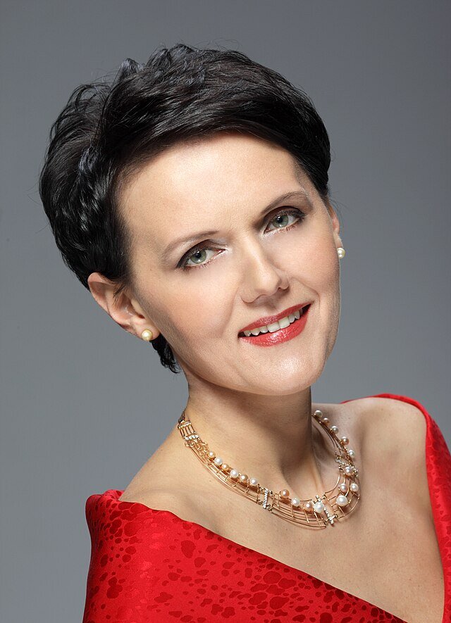 Portret uśmiechniętej kobiety z krótkimi ciemnymi włosami, w czerwonej sukni i perłowym naszyjniku, na neutralnym tle.