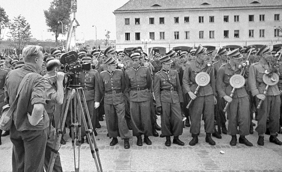 Zdjęcie przedstawia żołnierzy stojących w kilku równych rzędach, przed trzykondygnacyjnym budynkiem z dużą ilością okien. Mężczyźni ubrani są w jednolite mundury: kurtki, spodnie wpuszczone w buty do kostek, skórzane pasy, a  na głowach mają okrągłe czapki z otokiem i daszkiem. Kilku żołnierzy z pierwszego rzędu jest uzbrojonych w karabiny maszynowe. Przed zgromadzonymi, po lewej stronie,  stoi dwóch cywilów. Jeden z nich  filmuje zebranych przy użyciu kamery stojącej na trójnogu, drugi obserwuje sytuację, trzymając ręce na biodrach. 