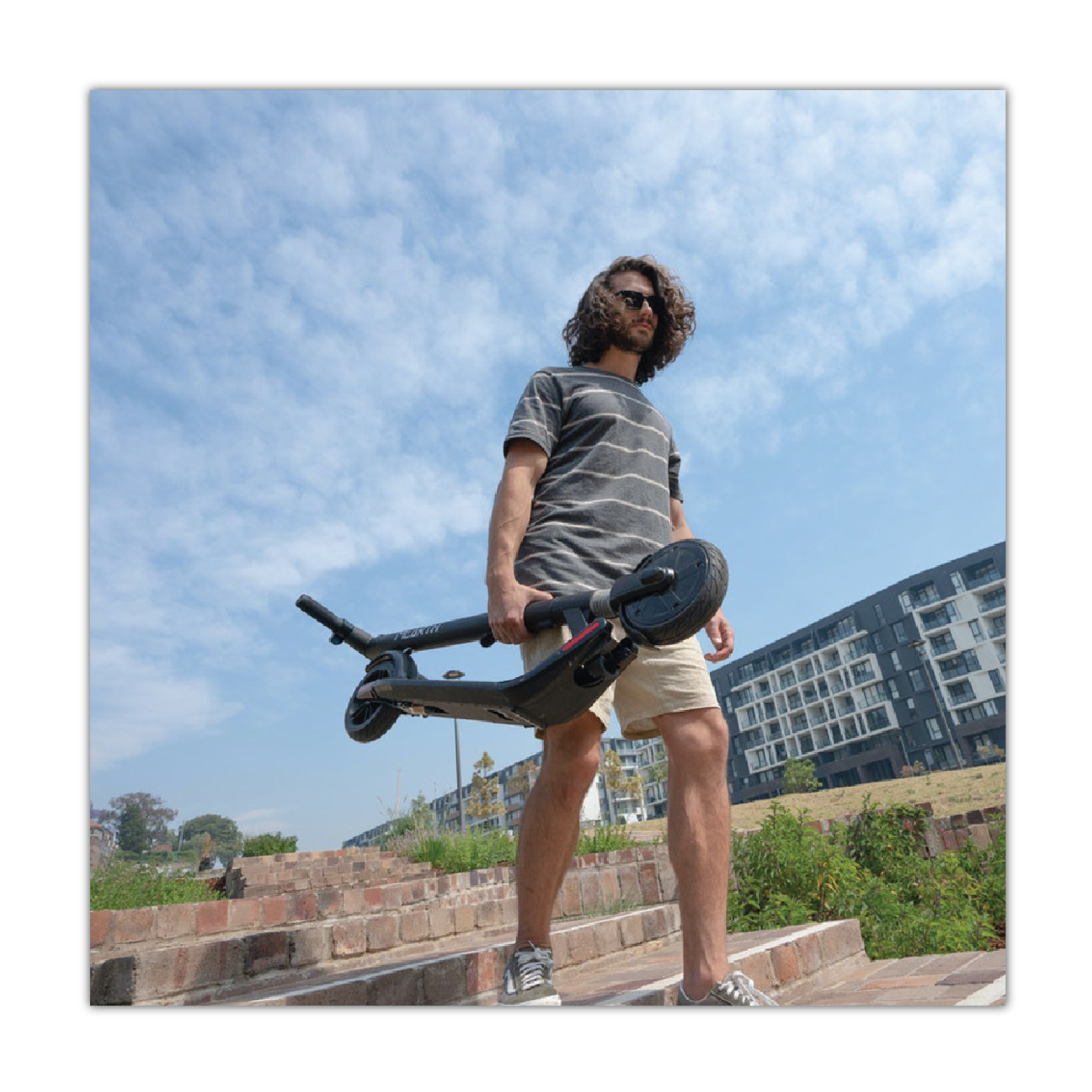 Na zdjęciu widoczny jest młody mężczyzna schodzący po schodach ze złożoną hulajnogą w prawej ręce. W tle blok mieszkalny.