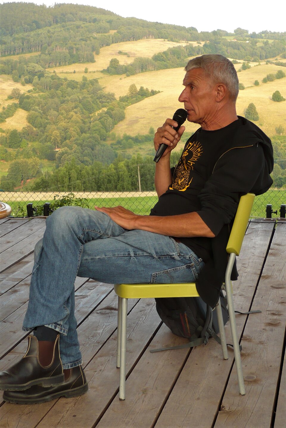 Zdjęcie przedstawia siedzącego na krześle dojrzałego mężczyznę. Siedzi bokiem, ma założoną nogę na nogę, mówi do mikrofonu. Ma krótkie, siwe włosy i zmarszczki na twarzy. Ubrany jest w dżinsowe spodnie oraz czarną koszulkę. W tle znajdują się wzniesienia z polami i lasem.
