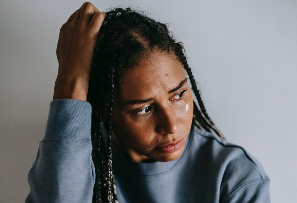 Zdjęcie przedstawia młodą, czarnoskórą kobietę. Po jej twarzy płyną łzy. Kobieta ma lekko schylona głowę, patrzy w swoja lewą stronę. Prawą dłonią podpiera głowę. Prawa część twarzy i nosa jest ocieniona. Za kobietą, na ścianie widoczny jest nieznaczny cień pozostawiony przez sylwetkę kobiety.