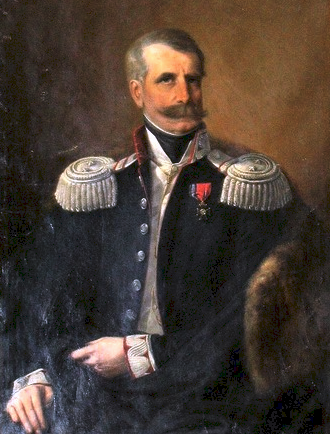 Ilustracja przedstawia mężczyznę w mundurze wojskowym. Ma na mundurze przypięty medal. Mężczyzna ma krótkie, zaczesane na bok włosy. Ma gęste wąsy oraz podłużną twarz.