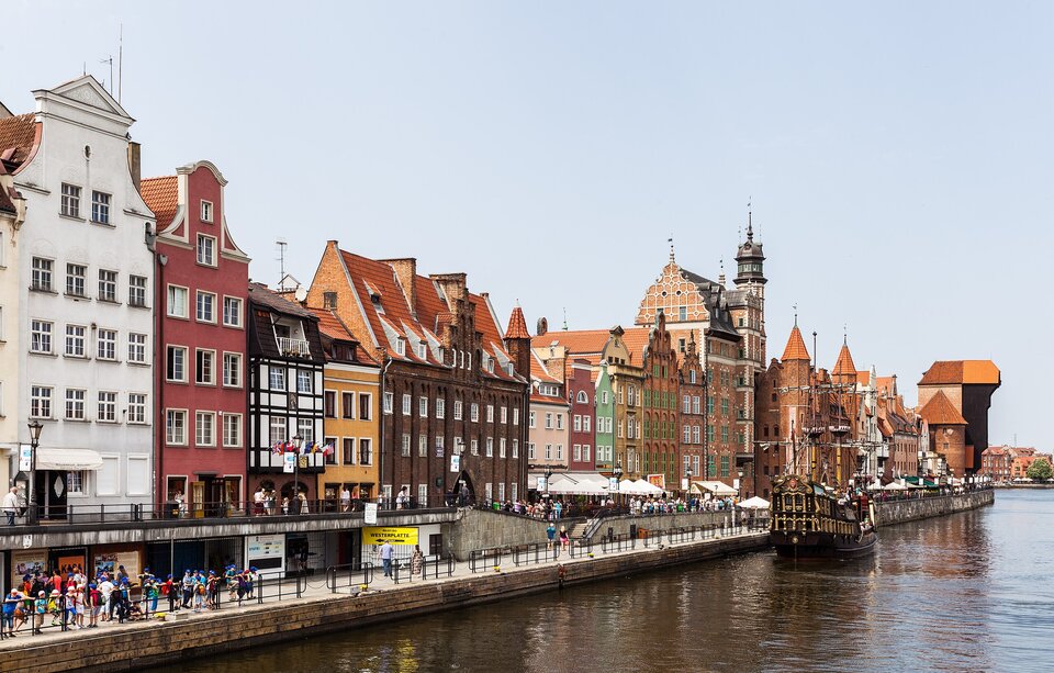 Widok historycznego nabrzeża nad rzeką w dużym mieście portowym. Po lewej stronie stoją ceglane kamienice z czerwonymi dachami oraz zabytkowy żaglowiec zacumowany przy deptaku pełnym ludzi i ogródków kawiarnianych, dalej w głębi dominuje masywna średniowieczna brama–żuraw portowy, a po prawej stronie widać nowoczesny most nad wodą, zabudowę śródmiejską i fragment portu.