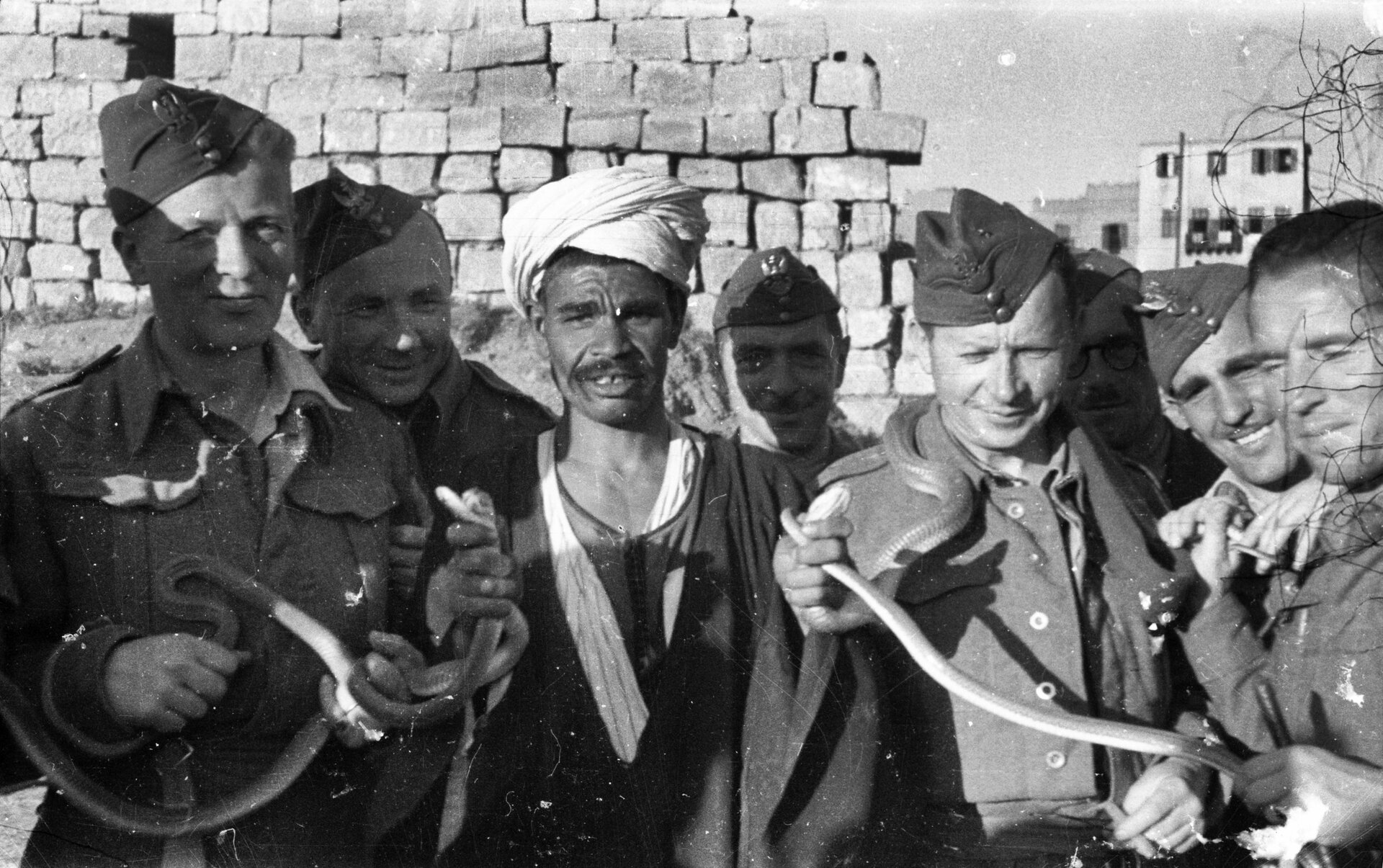 Zdjęcie przedstawia mężczyznę w turbanie na głowie, otoczonego uśmiechniętymi, polskimi żołnierzami. Żołnierze trzymają w dłoniach kilka węży.