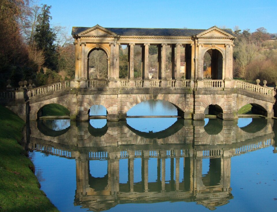 Fotografia przedstawia staw w parku. Nad wodą wznosi się kamienny most. Zadaszenie opiera się na ozdobnych kolumnach. Budowla odbija się w lustrze wody. Wokół rosną drzewa. Niebo jest bezchmurne.