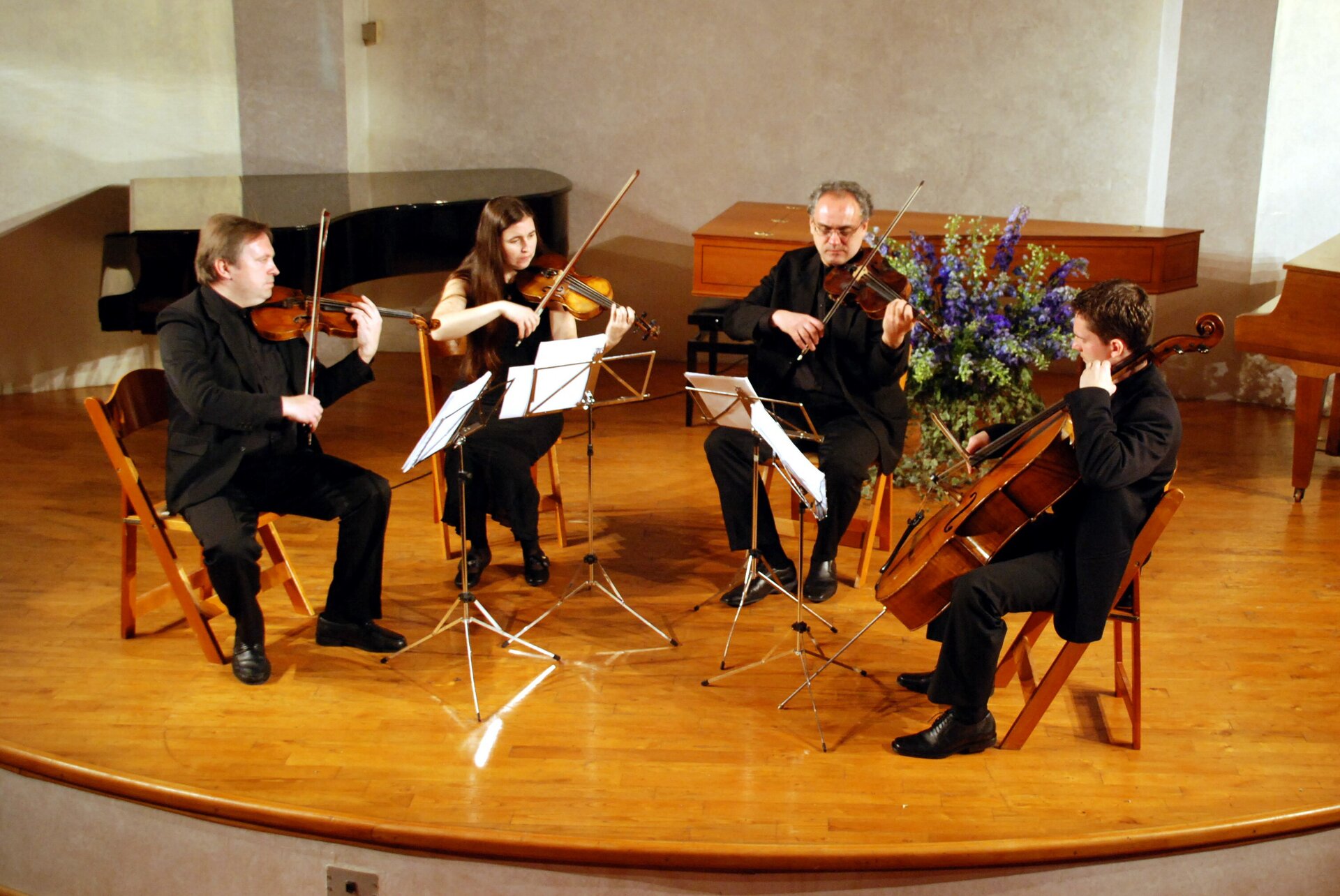 Zdjęcie przedstawia kwartet smyczkowy. Na scenie znajdują się cztery osoby siedzące obok siebie, ubrane w galowe stroje. Każda z osób gra na innym instrumencie: od lewej do prawej: pierwsze skrzypce, drugie skrzypce, altówka oraz wiolonczela. W tle znajdują się trzy pianina.