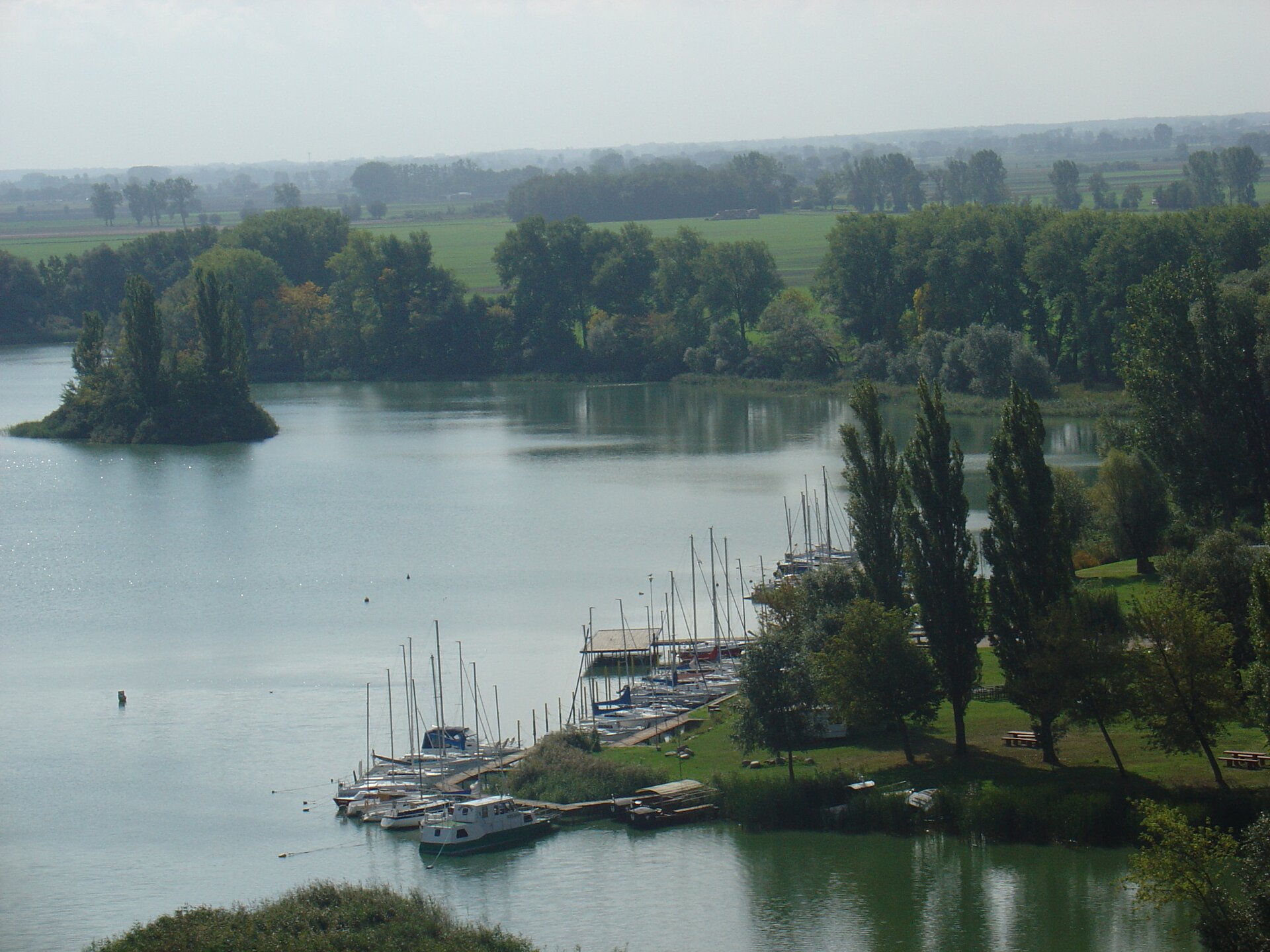 Kolorowa fotografia przedstawia fragment jeziora Gopło widziany z lotu ptaka. Otaczają je lasy i łąki. Z prawej strony widać przystań dla jachtów. Stoją tu zacumowane jachty z opuszczonymi żaglami.